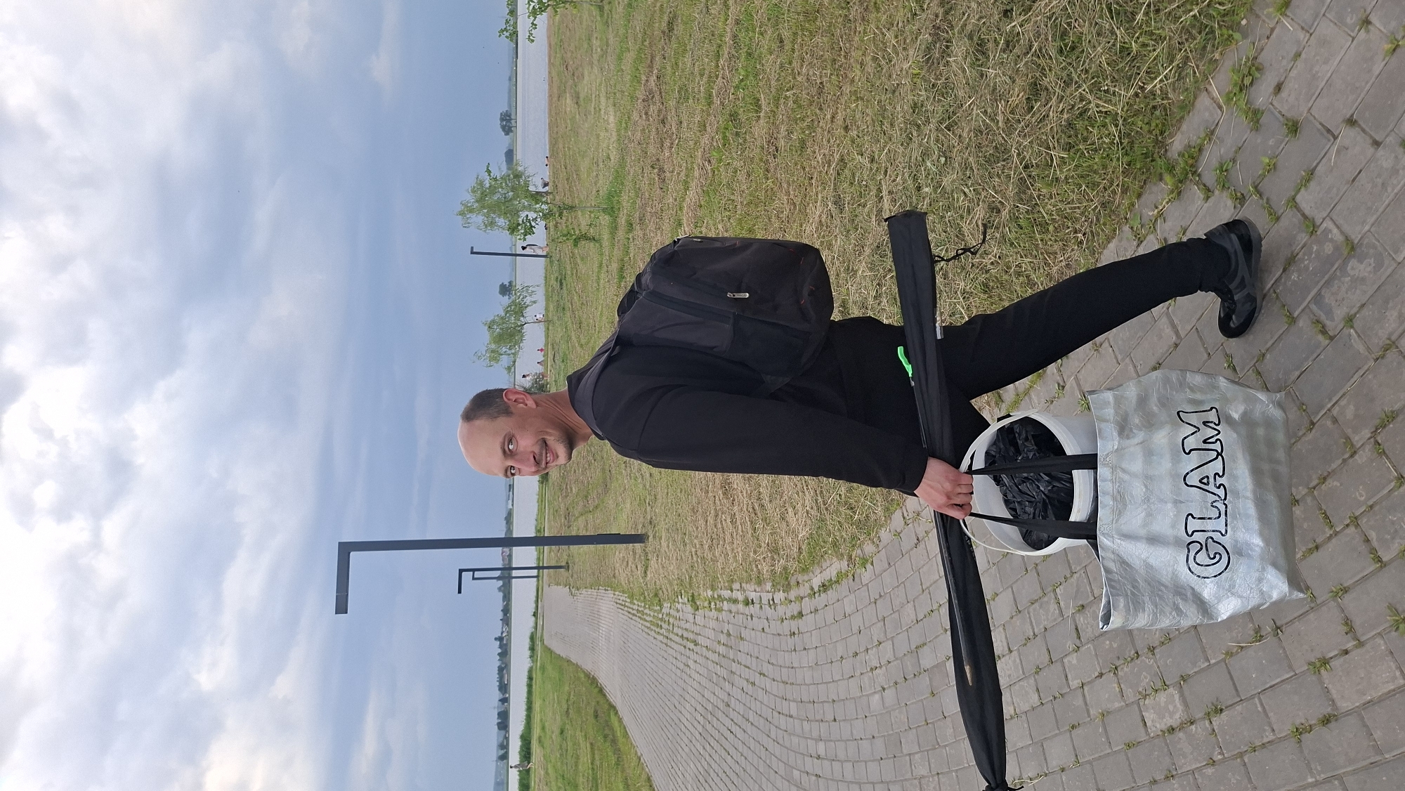 A bald man wearing a black sweatshirt and black pants, with a black backpack on his back, walks along a paved pathway next to a grassy area. He is carrying a white bucket with black plastic bags inside and a long black case, possibly for fishing rods, held horizontally across his front. In the background, a body of water is visible, with trees and a few people in the distance on the far shore. The sky is overcast. The pathway appears to be in a park or recreational area, possibly near the Smoljavicskaje reservoir in Smoljavičy, Belarus, given the presence of a body of water and recreational features.