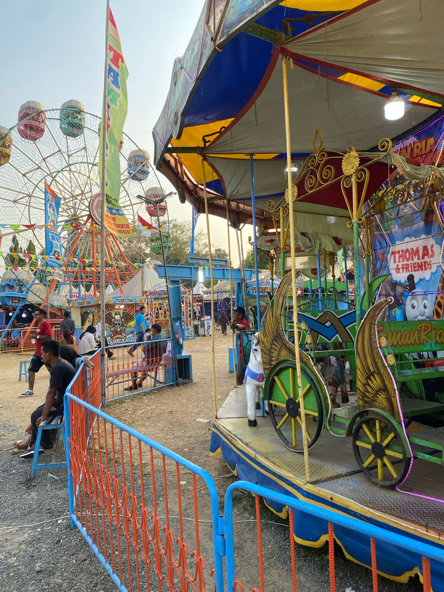 The image depicts a bustling outdoor carnival scene, likely located in Nyamok, Indonesia, with a bright, sunny sky overhead. In the foreground, a colorful carousel, decorated with images of characters from "Thomas & Friends," features whimsical winged horses and green carriages with yellow wheels. A blue and orange barrier separates the viewer from the carousel area. Beyond the carousel, a large Ferris wheel with multi-colored passenger cabins dominates the mid-ground, surrounded by various carnival tents and structures. Several people are visible throughout the scene, some seated on benches or standing, while others appear to be patrons or workers at the carnival. The ground is covered with a mixture of dirt, gravel, and loose stones, indicative of a temporary outdoor event. The overall atmosphere suggests a lively and festive environment.