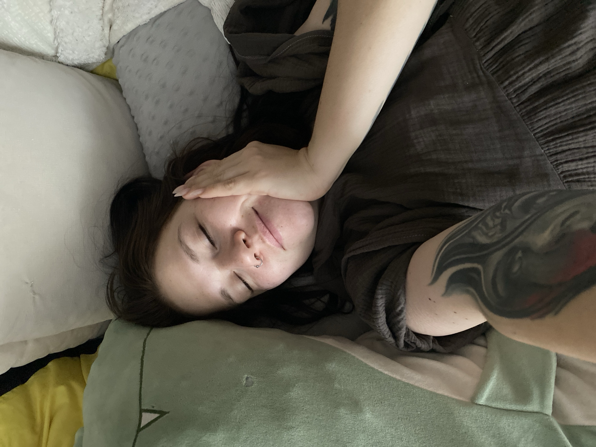 A young woman lies on a bed. She is turned to her left and covering her face with her right hand. She has a nose piercing on her left side. The woman is wearing a gray shirt or dress. Her left arm is visible, and it has a dark-colored tattoo. Her head rests on a green plush pillow with a triangle pattern, which is next to a white pillow and a yellow object. There is also a gray pillow in the background. This scene is within an indoor setting, presumably a bedroom. There is no information on the exact location of this image being in the city of Chaikovsky, Russia, however.