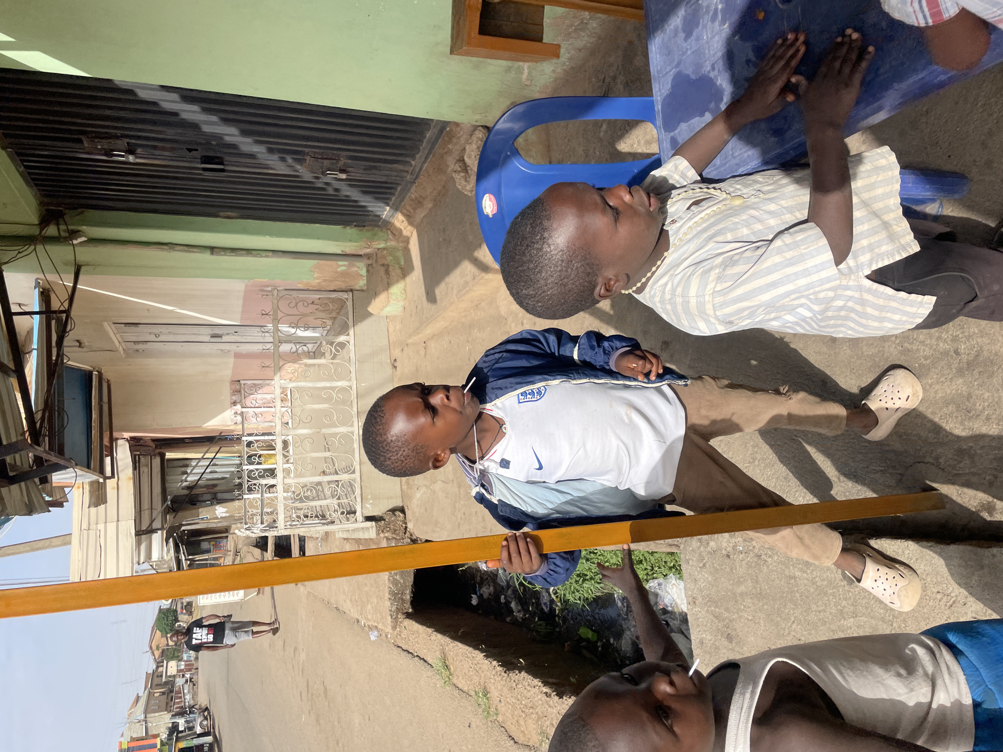 Two young boys and partial view of a third are visible in an outdoor setting. One boy holds a yellow rectangular object. Another is seated at a blue plastic table. Some objects include chairs, a street, and buildings. The location is likely Jos, Nigeria.