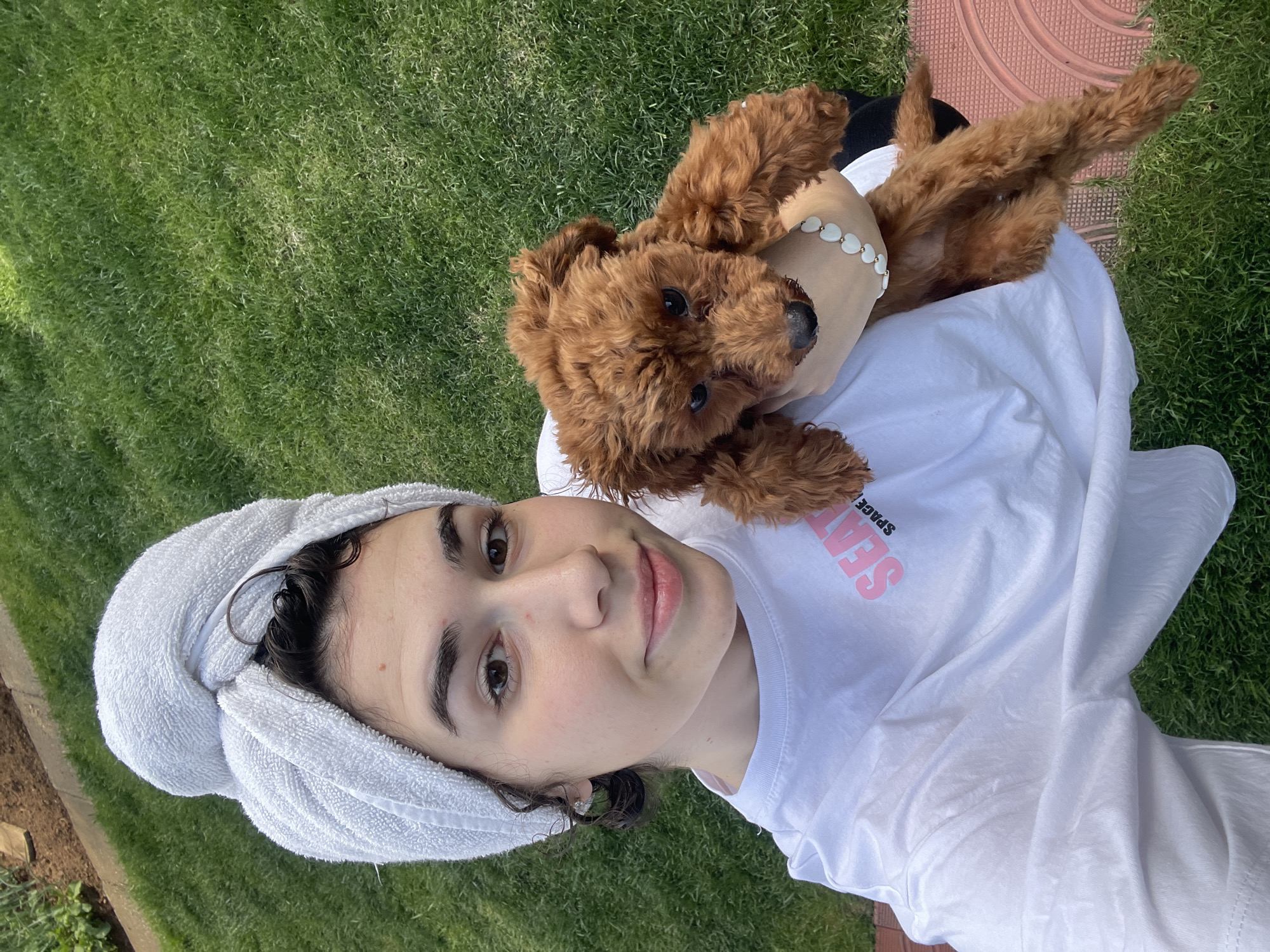 A young woman with a towel wrapped around her head poses for a selfie with a brown curly-haired dog in a grassy outdoor setting. She is wearing a white t-shirt with pink lettering. The dog is nestled on her chest, with its legs outstretched. The background includes green grass and a section of red-toned ground cover. The location is unidentifiable as Sofia, Bulgaria based on the visible information.