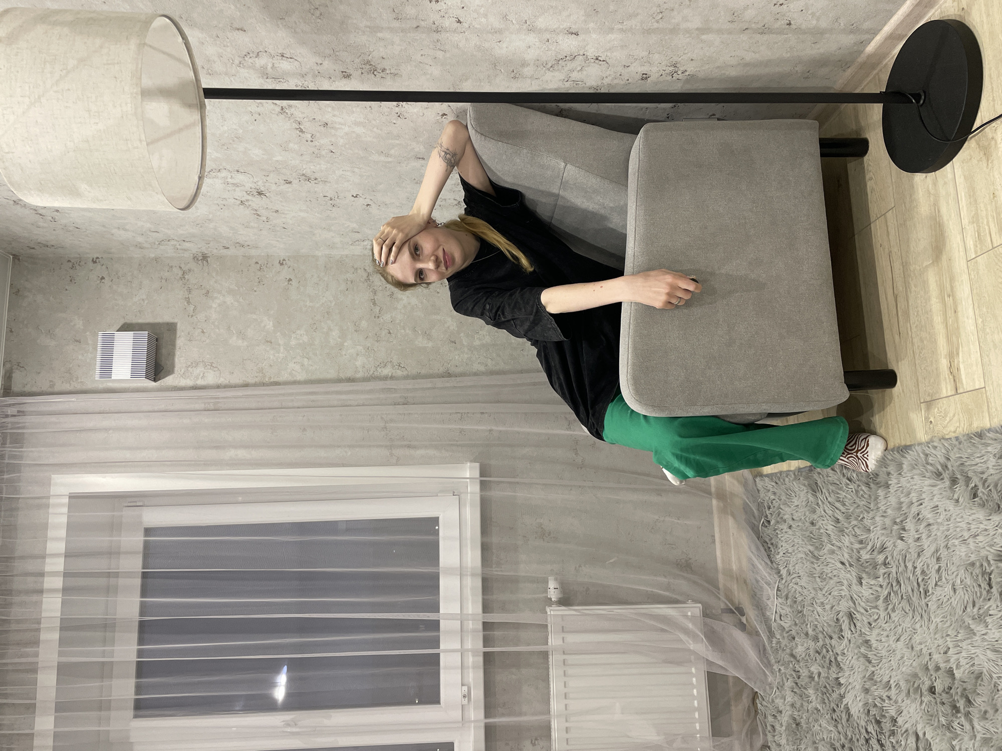A young adult woman is reclining on a grey loveseat, appearing to pose for the camera. She has blonde hair pulled back in a ponytail, wears a black long-sleeved shirt, and green pants. Her right hand rests on her head, and her left hand is on the arm of the loveseat. A floor lamp stands behind the loveseat, with a white drum shade above and a black circular base on the floor. A window with sheer white curtains is visible to the left, with a radiator below the window. The walls are light grey with a textured pattern, and the floor is a mix of light-colored wood and a fluffy grey rug. A small white air conditioner unit is mounted high on the wall to the left of the window. The location is likely a residential interior in Izhevsk, Russia, based on the photographic style and typical furnishings seen in the region.