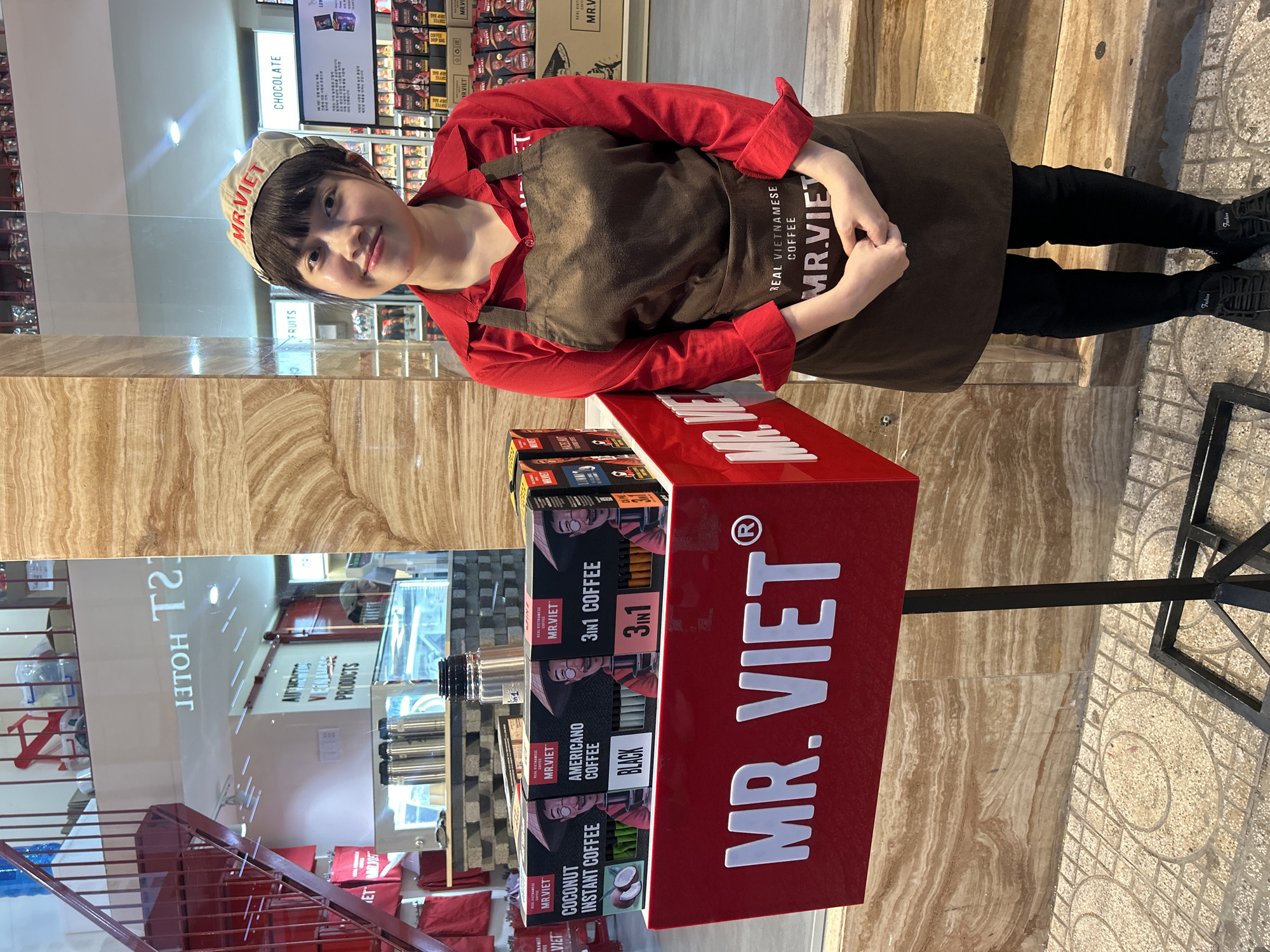The image captures a scene at a Mr. Viet coffee retail location, likely in Nha Trang, Vietnam. A young woman wearing a red shirt, dark brown apron with "MR. VIET REAL VIETNAMESE COFFEE" printed on it, and a tan cap with "MR. VIET" embroidered on it stands next to a red display stand. The display stand features the "MR. VIET" logo prominently and holds boxes of Mr. Viet instant coffee in various flavors, including Coconut, Americano Black, and 3in1 coffee. Behind her is a store with various products on display, and a reflection indicates that the store may be located in a hotel or shopping center. The floor transitions from an interior tiled surface to an exterior tiled pavement with circular patterns, indicating a transition from indoor to outdoor space.