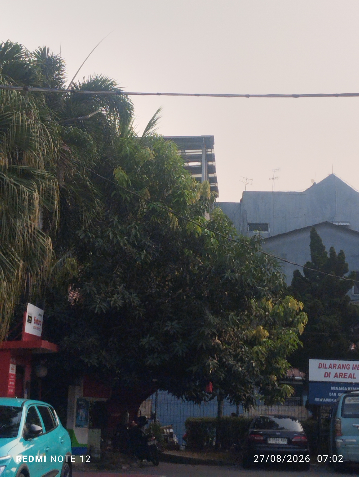 A low-angle, medium shot captures a tree-lined street in Bekasi, Indonesia, at dusk. The foreground is dominated by the lush green canopy of several trees, with palm fronds from the left extending into the frame. Partially visible is a turquoise car on the left, with the text "REDMI NOTE 12" imprinted on its surface. Behind the trees, a red building with a white sign bearing the word "Enduro" is discernible. Further in the background, a series of blue-grey buildings with antennas on their roofs suggest a residential or commercial area. To the right, a white sign with red text reads "DILARANG MEROKOK DI AREAL", indicating a no-smoking zone. The sky is overcast, with the sun seemingly set or obscured by clouds, casting a soft, diffused light on the scene. A date and time stamp "27/08/2026 07:02" is present at the bottom right of the image.