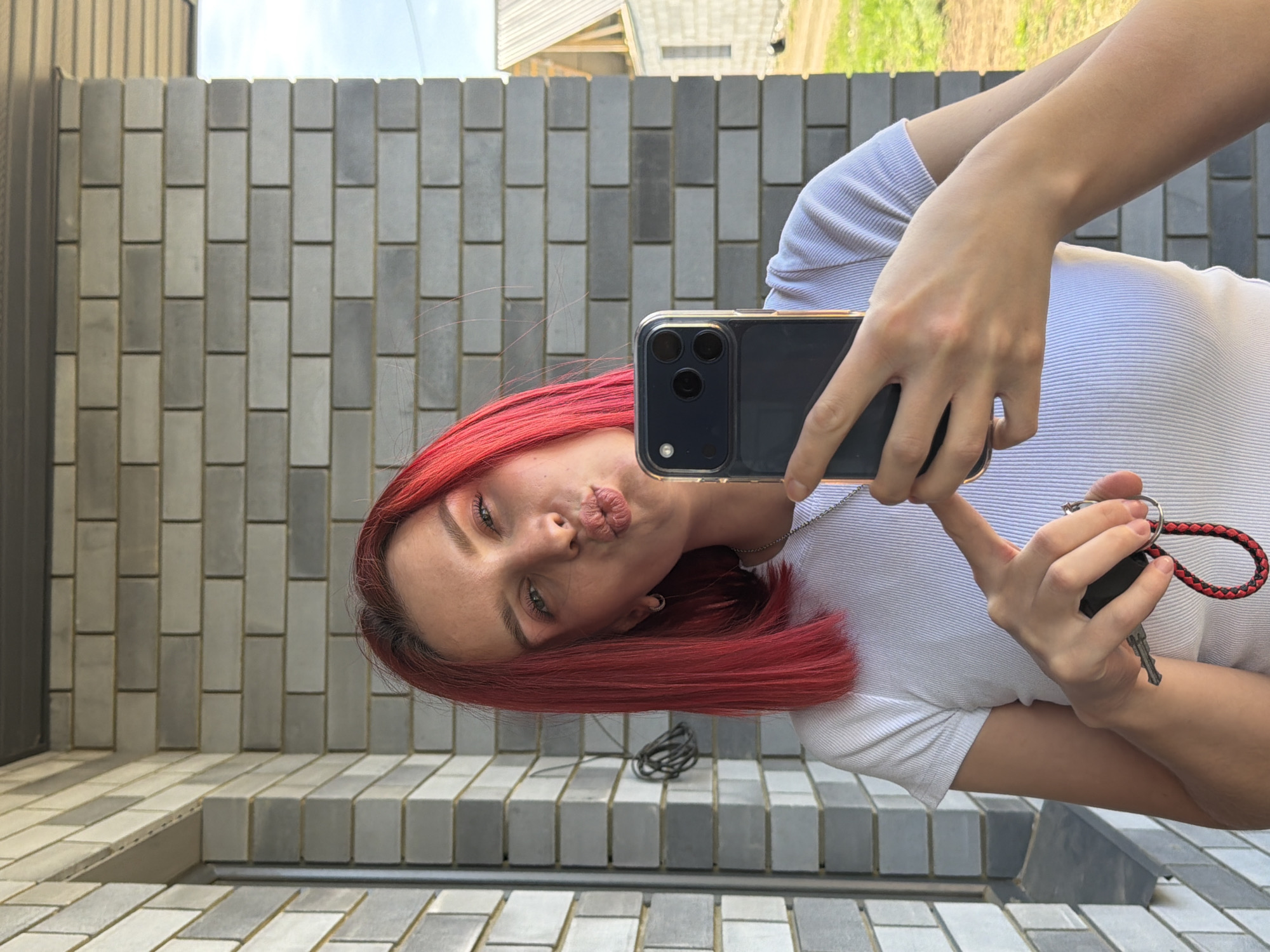 A young woman with red hair is taking a selfie in what appears to be an outdoor setting in Chaikovsky, Russia. She is holding a cell phone in one hand and a set of keys in the other, with the keys having a red and black braided lanyard. The woman is wearing a white, ribbed t-shirt. The background is a wall constructed of gray bricks, likely a fence or the side of a building. A glimpse of a building and greenery is seen above the wall in the background, suggesting an urban or suburban residential area.