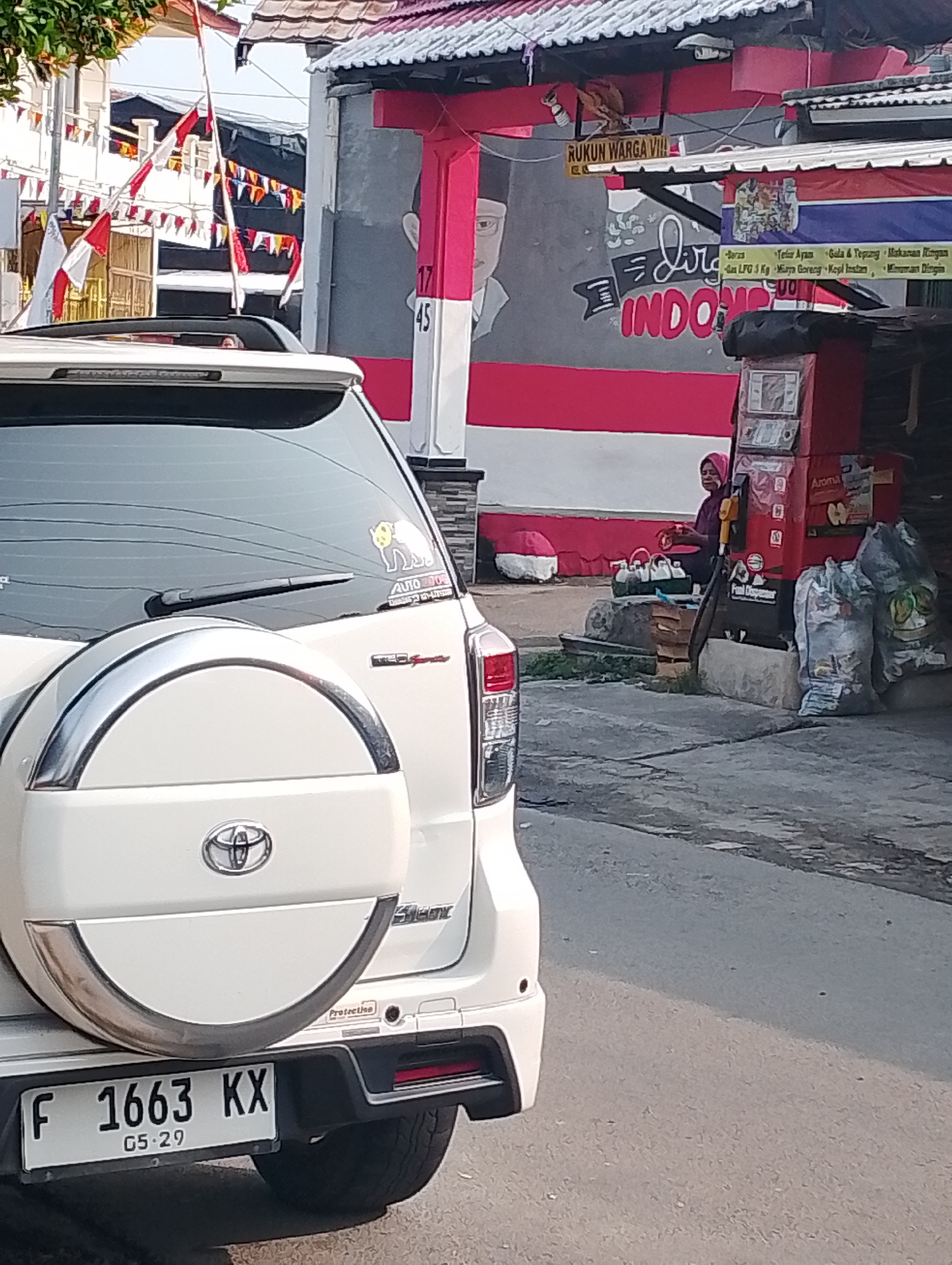 A white SUV is parked on a paved street, its rear facing the camera. The SUV has a spare tire mounted on the back. In the background, a building is decorated with red and white Indonesian flags. A mural on the building depicts a portrait of a man and the words "DIRGAHAYU INDONESIA." A woman in a pink headscarf is sitting near a small stall filled with various items. The scene appears to be in Bogor, Indonesia, given the presence of Indonesian flags and the text on the mural.