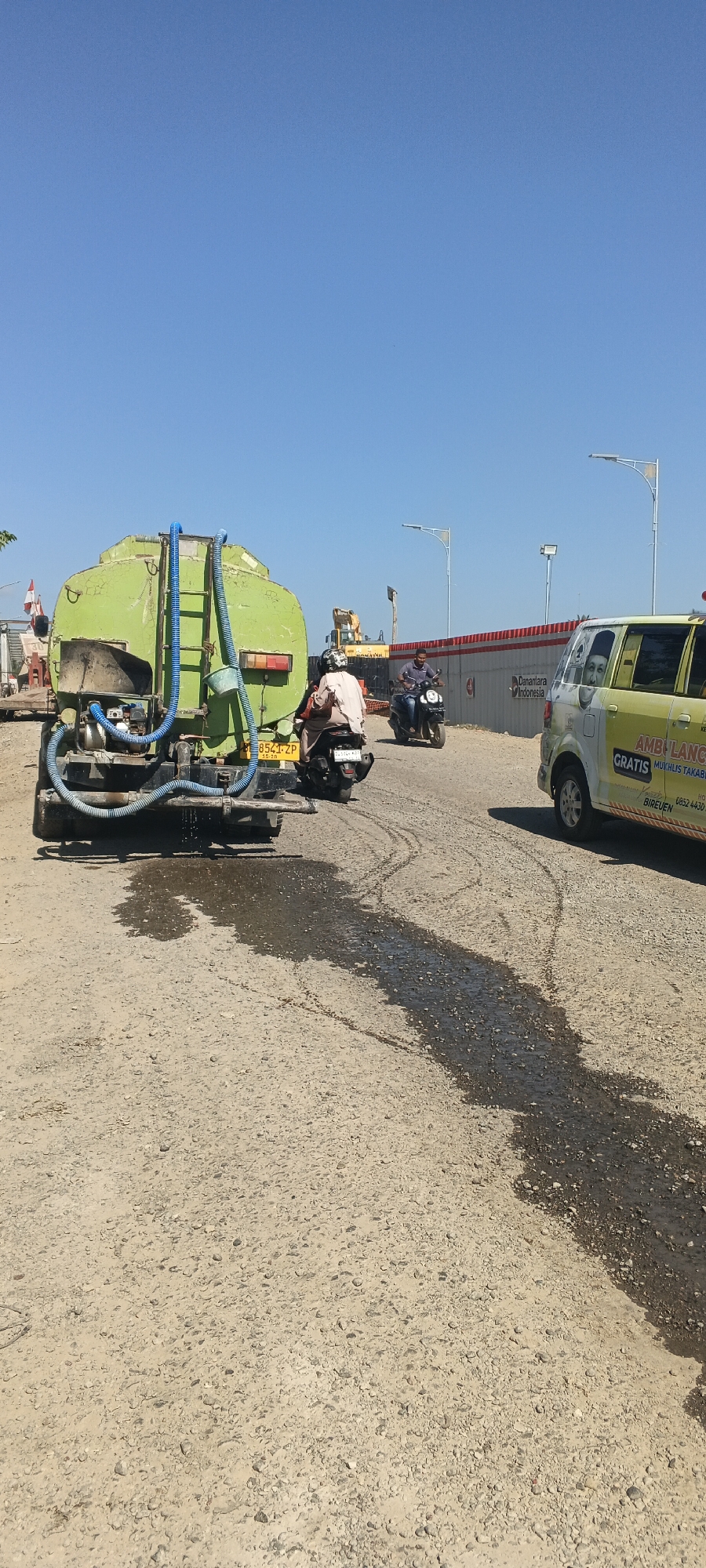 In Blang Mee, Indonesia, a scene unfolds on a dusty, unpaved road under a clear blue sky. A large, green water truck, likely involved in road maintenance, is positioned on the left, with water spraying from its rear onto the road surface, creating a wet patch. A person on a motorcycle is visible in front of the truck, facing away from the camera. Further down the road, another person on a motorcycle is moving away from the viewer. To the right, a yellow van, an ambulance, is parked or moving, displaying text that includes "AMBULANCE GRATIS" and "MUKHLIS TAKABUL" with a phone number. The background suggests ongoing construction, with a yellow excavator and a red and white barrier visible. Streetlights are also present along the road. The overall impression is of a work-in-progress environment with active roadwork and transportation.