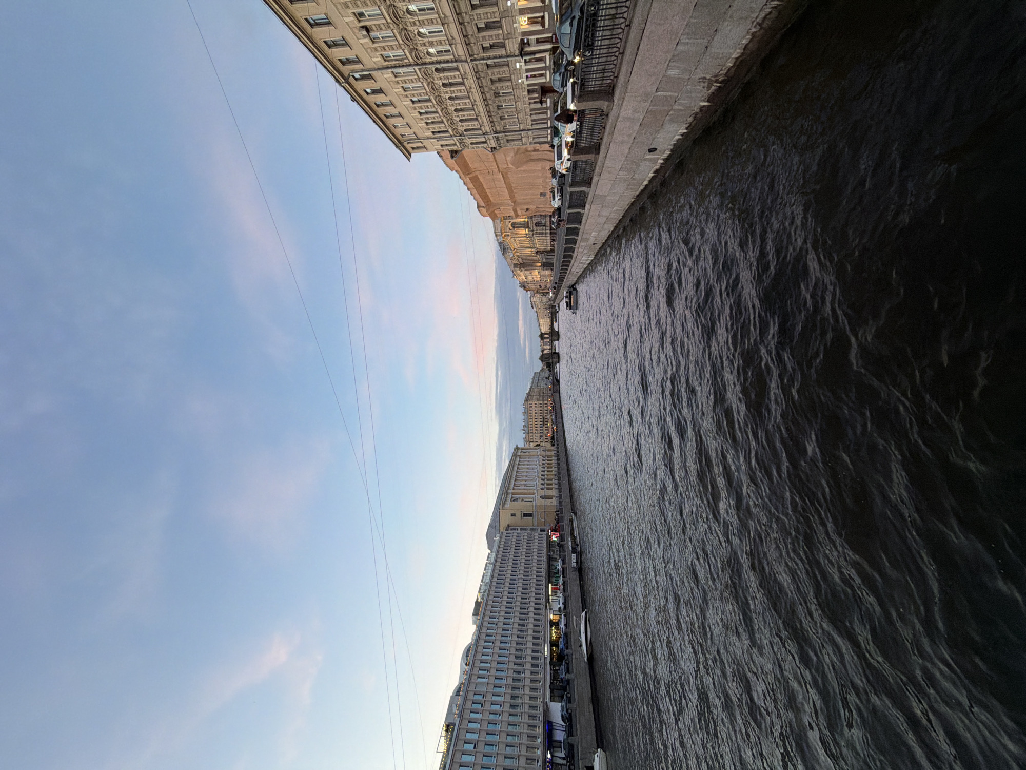 The image depicts a panoramic view of the Griboyedov Canal in Saint Petersburg, Russia, during twilight. The canal, a dark, rippling body of water, occupies the lower half of the frame. Along the left bank, a long, modern building with numerous windows and a lit facade is visible, with cars parked along the street in front. Further down the canal, older, classical-style buildings with ornate architecture line the right bank, some illuminated and others in shadow. Several boats are moored along the canal, and a few vehicles are parked on the embankment. The sky above is a soft gradient of blue and pink, with thin clouds and visible electrical wires stretching across the frame. The overall scene suggests an urban setting at dusk, showcasing the architectural contrast between older and newer structures along the waterway.