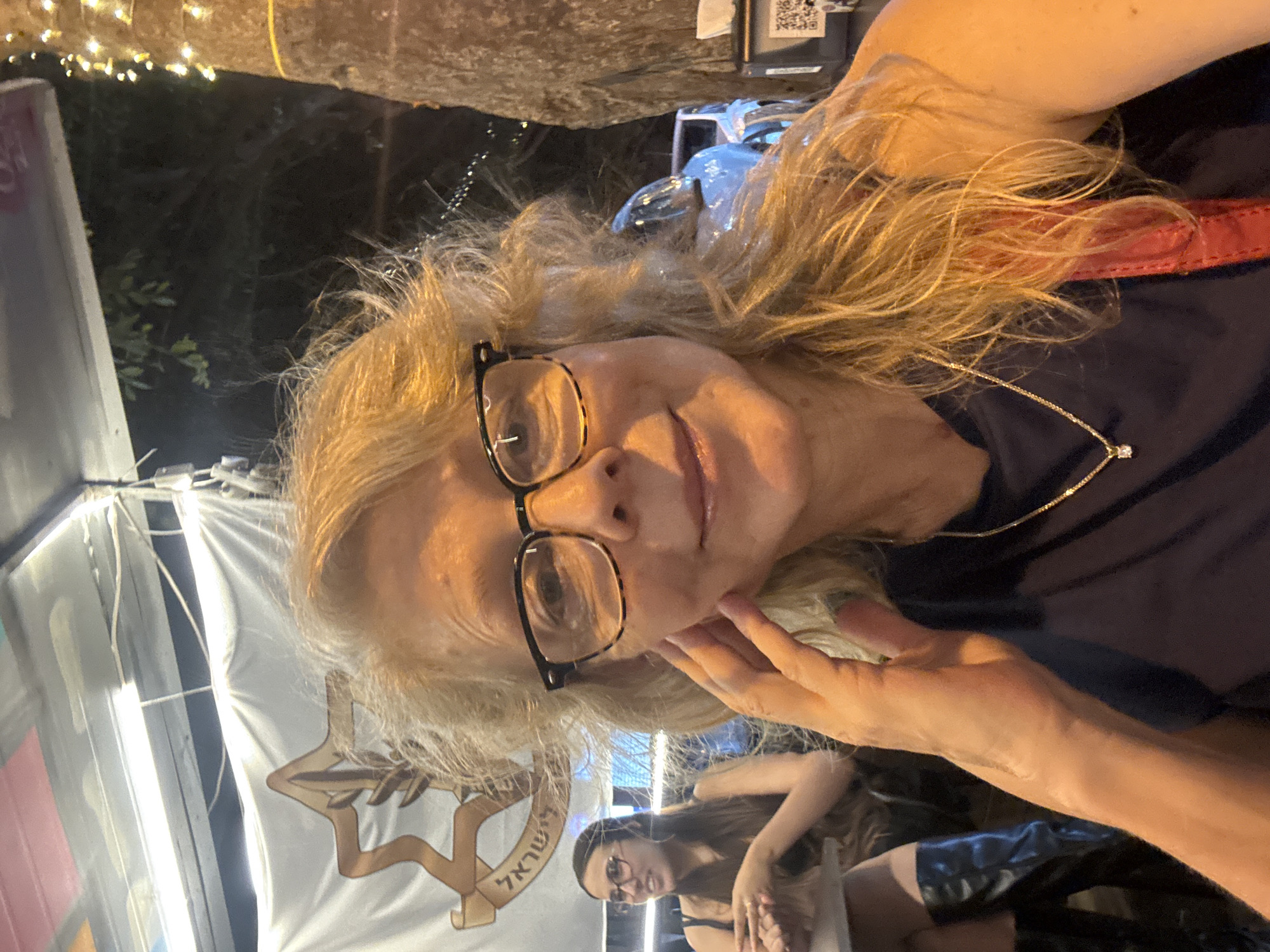 The image is a selfie taken outdoors at night, likely in Ramat Gan, Israel, given the Israeli flag visible in the background. A woman with blonde, wavy hair and glasses is in the foreground, looking at the camera. She is wearing a dark top and a necklace. Her right hand is raised to her cheek. In the background, another woman with dark hair and glasses is seated at a table, talking to someone out of frame. The scene appears to be an outdoor dining or seating area, with a tree decorated with lights to the right of the frame. There are also cars and what looks like a building in the background.