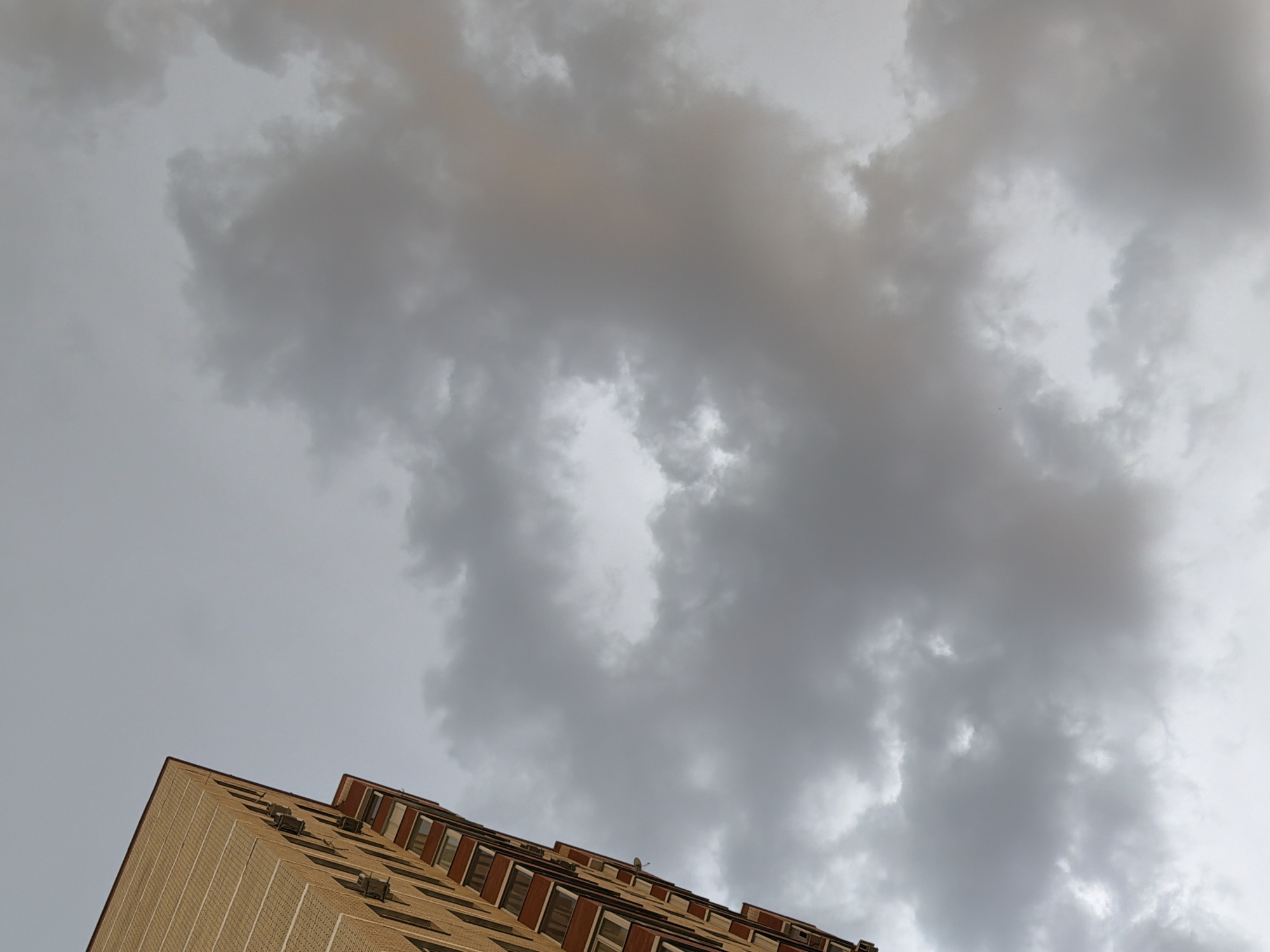 The image shows a low-angle view of the top portion of a tall, multi-story building against a backdrop of heavy, gray storm clouds. The building appears to be constructed of light-colored brick with dark window frames. There are protrusions from the facade, possibly air conditioning units or vents. The location appears to be in a city, though the specific city is Unknown, Unknown. The primary subjects are the building and the clouds, with no people visible. The environment suggests an overcast or stormy day.