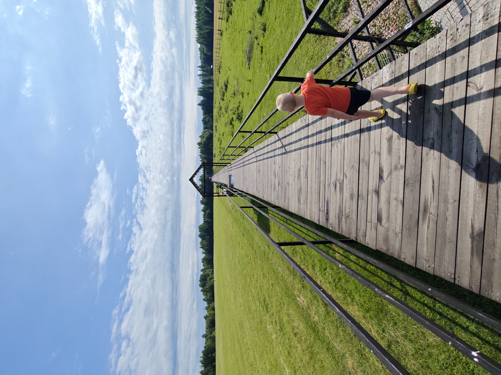 A young boy with blond hair, wearing an orange t-shirt and dark shorts, walks on a wooden pier that extends into a vast, bright green, mowed field. The pier has black metal railings. In the distance, the pier leads to a covered structure, possibly a gazebo. A line of trees forms the horizon behind the field, and a fence runs along the edge of the field. The sky above is a clear blue with scattered white clouds. The scene is set in Dorishchi, Russia, on a sunny day. The boy's shadow is cast long on the wooden planks of the pier.