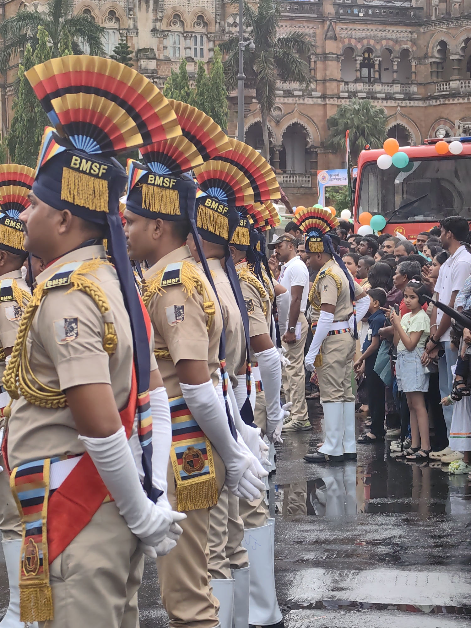 The image depicts a ceremonial procession in front of the Chhatrapati Shivaji Maharaj Terminus (CSMT) in Mumbai, India. Several individuals in uniform, adorned with traditional Bawa, are marching, their distinctive headdresses and sashes prominent. The uniforms are beige with blue and red accents. Behind them, a crowd of spectators lines the wet street, observing the event. A red vehicle, possibly a fire truck, decorated with balloons in white, orange, and green, is visible in the background, along with palm trees and the historic architecture of CSMT. The ground appears wet, indicating recent rain.