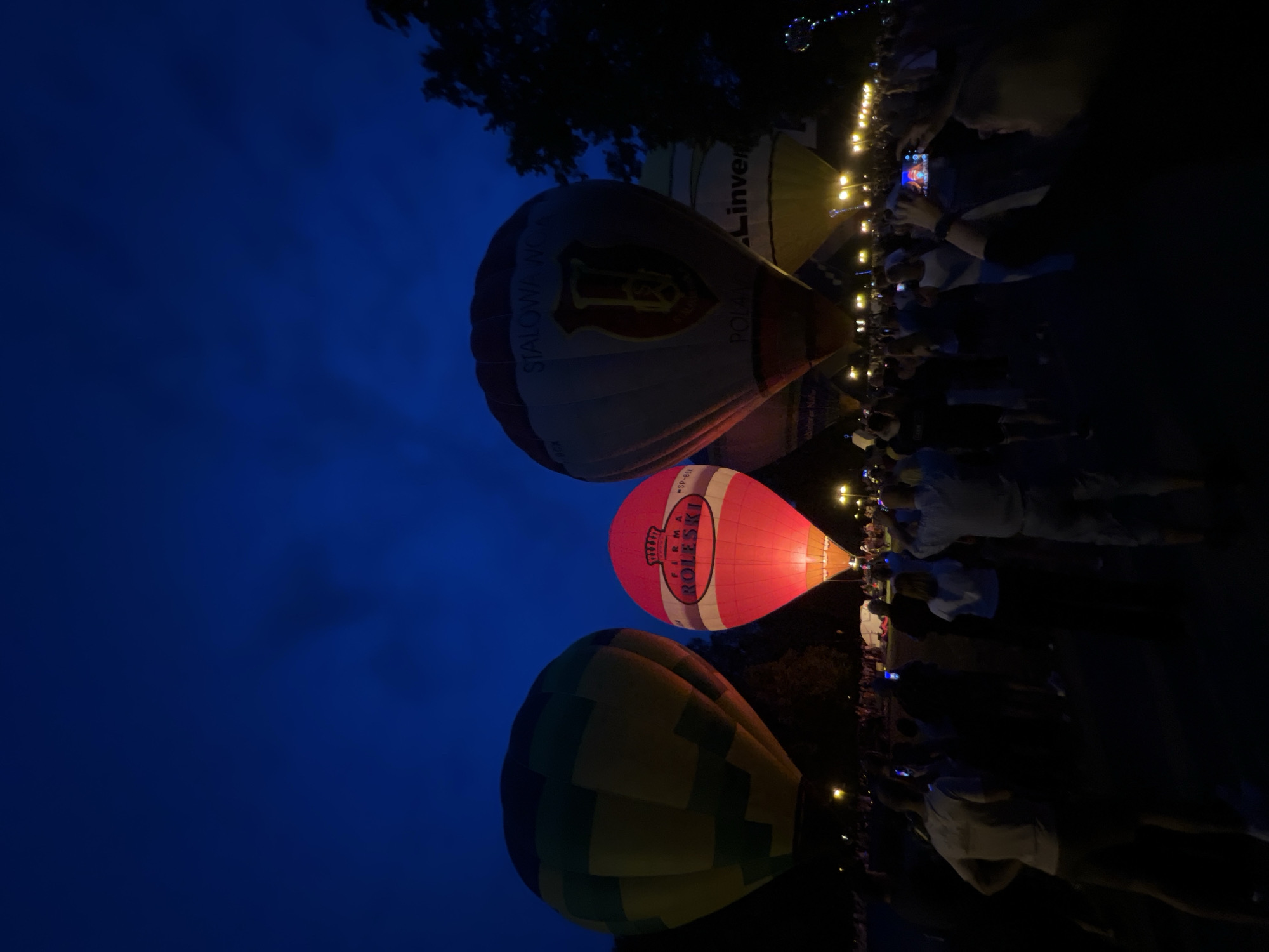 The image shows a night-time event featuring hot air balloons in Tychy, Poland. Several hot air balloons are visible against a dark blue, twilight sky. One prominent balloon is bright pink with a white oval logo containing red text that reads "FIRMA ROKESKI". Another balloon is a lighter color, possibly white and maroon, with a crest-like logo and text on its side. Further in the background, a yellow and white balloon is partially visible with the word "INVER" clearly legible. A green and yellow checkered balloon is in the foreground.
A crowd of people is gathered, many holding up their phones to capture the spectacle, suggesting a festival or public viewing. The scene is illuminated by the glow of the balloons and string lights that line what appears to be a pathway or perimeter. The overall atmosphere seems festive and engaging, with people captivated by the illuminated balloons. The time of day is clearly evening or night, as indicated by the dark sky and artificial lighting.