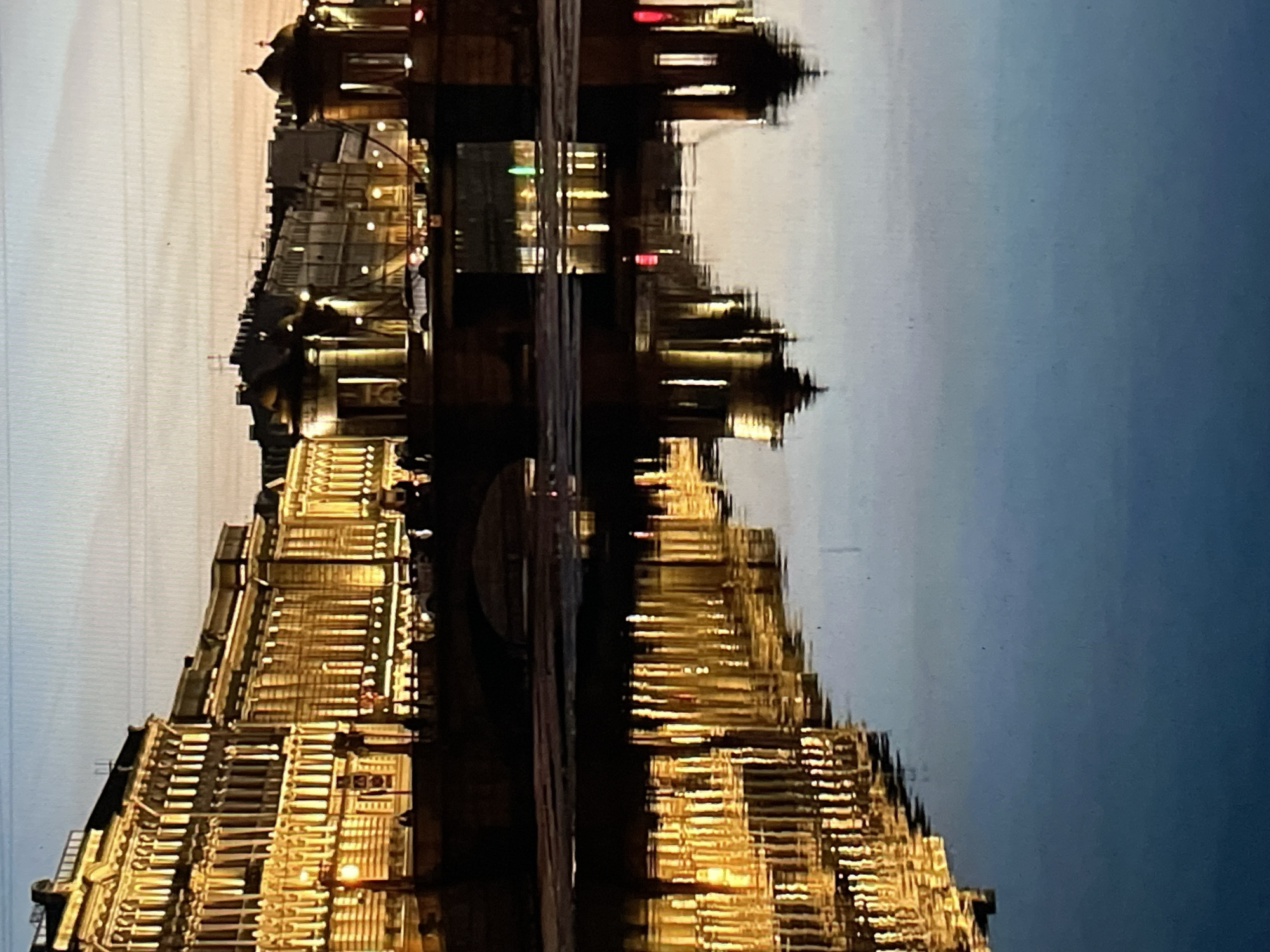 The image captures a twilight or nighttime view of the Lomonosov Bridge over the Fontanka River in Saint Petersburg, Russia. The left bank features a row of multi-story neoclassical buildings, uniformly illuminated with warm yellow lighting, emphasizing their facades, windows, and architectural details. The Lomonosov Bridge, a multi-arch stone structure, crosses the river, with two distinctive, illuminated domed pavilions visible on its right side. Several indistinct human figures are silhouetted against the bridge's railing and illuminated background, appearing to walk. The calm, dark blue surface of the Fontanka River occupies the lower half of the frame, providing clear reflections of the illuminated buildings, bridge pavilions, and various light sources, including a green light reflection near the bridge's center. Subtle ripples distort the reflections slightly. The sky transitions from dark blue overhead to a light orange-pink glow at the horizon. Distant city buildings and streetlights are visible beyond the bridge in the background. Patches of darker material are present on the water surface in the mid-ground.
