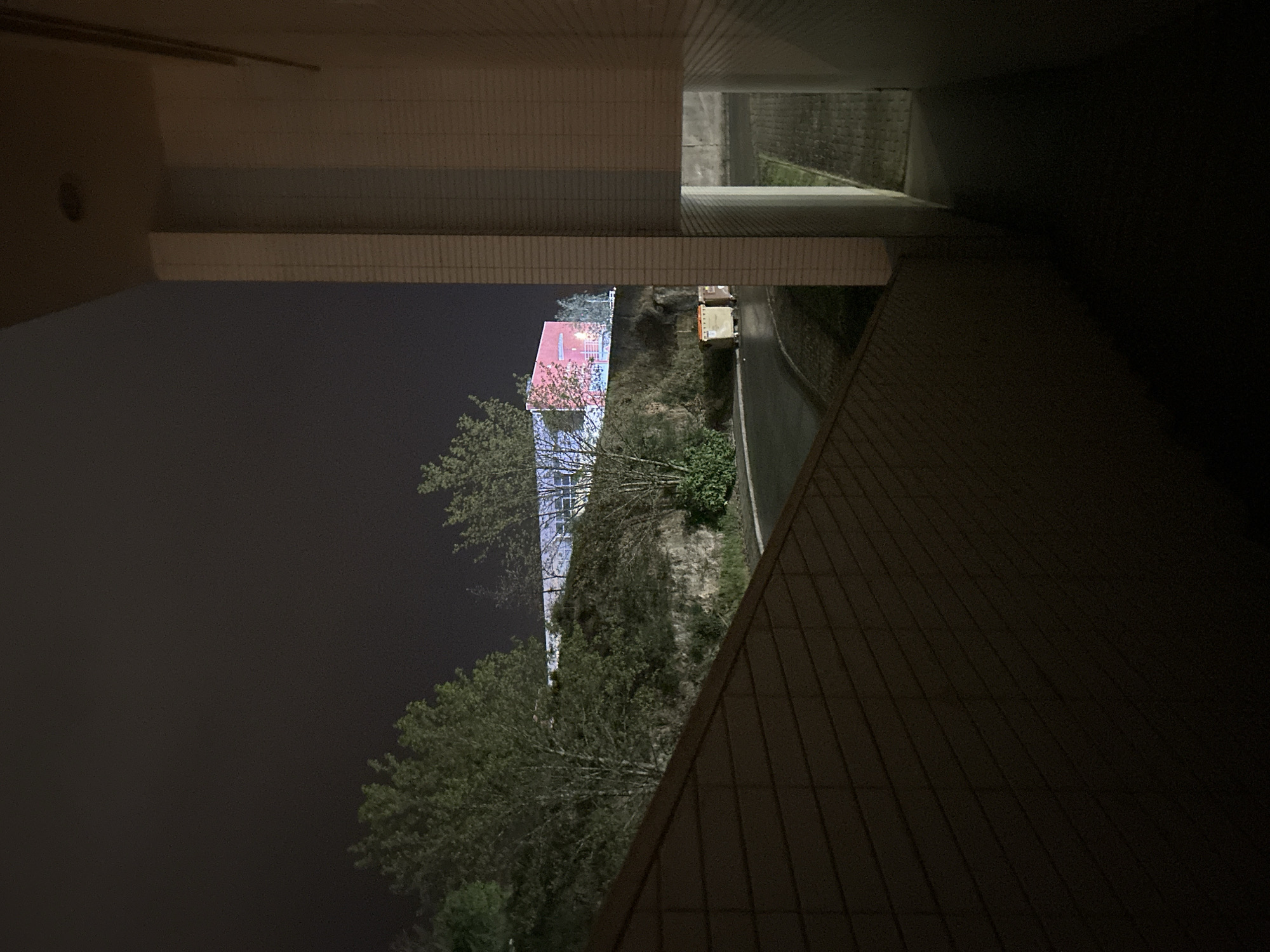 The image shows a building exterior at night, possibly in Vila Real, Portugal, based on architectural style and vegetation. The perspective is from under an overhanging structure with tile accents. In the background, a multi-story building with pink and white elements is visible amidst trees and vegetation. A street is also discernible, with a vehicle parked alongside it. The surroundings appear to be residential or mixed-use. The lighting is dim, emphasizing the darkness of the night.