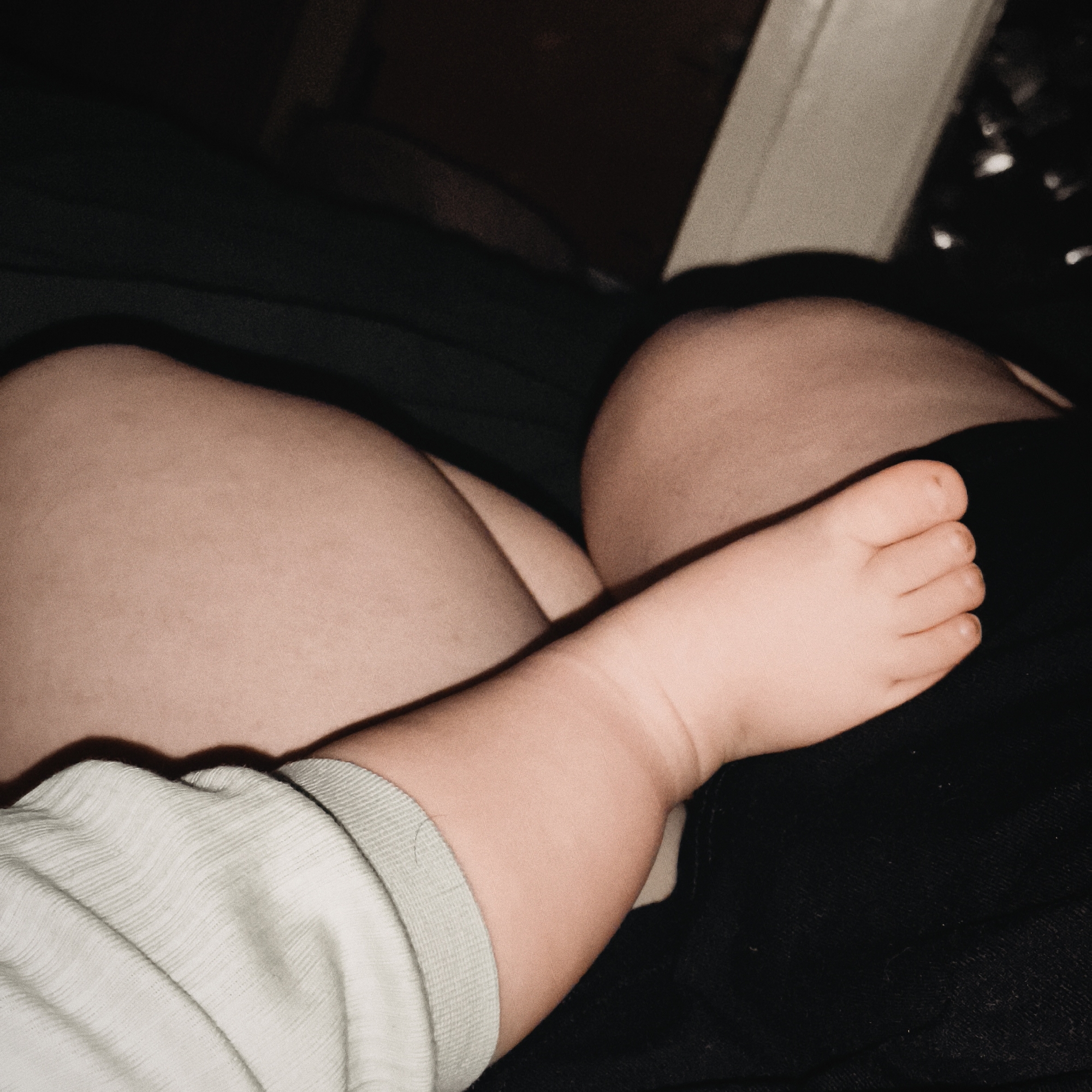 The image presents a close-up, intimate scene centered on the limbs of an adult and a baby or young child. In the foreground, the child's bare foot, with its tiny toes, rests gently on a dark fabric, likely the clothing of an adult. The child's arm, clad in a light green, possibly ribbed or textured fabric, is also visible on the left side of the frame.
The background is dominated by the adult's bare thighs or legs, exhibiting warm, natural skin tones. These body parts are softly out of focus, creating a comforting, enveloping space for the child. The adult's legs appear bent, suggesting a relaxed posture, perhaps lying down on a bed or sofa. Dark fabrics and shadows fill parts of the background, enhancing the sense of a dimly lit, private indoor setting.
The lighting is low and warm, implying either evening, nighttime, or a softly illuminated room, contributing to an atmosphere of calm and tenderness. The primary activity appears to be resting or snuggling, highlighting the physical closeness between the child and adult. The overall composition conveys a feeling of peace, love, and protection. There are no distinct location cues within the image itself, beyond being indoors. No visible text is present.