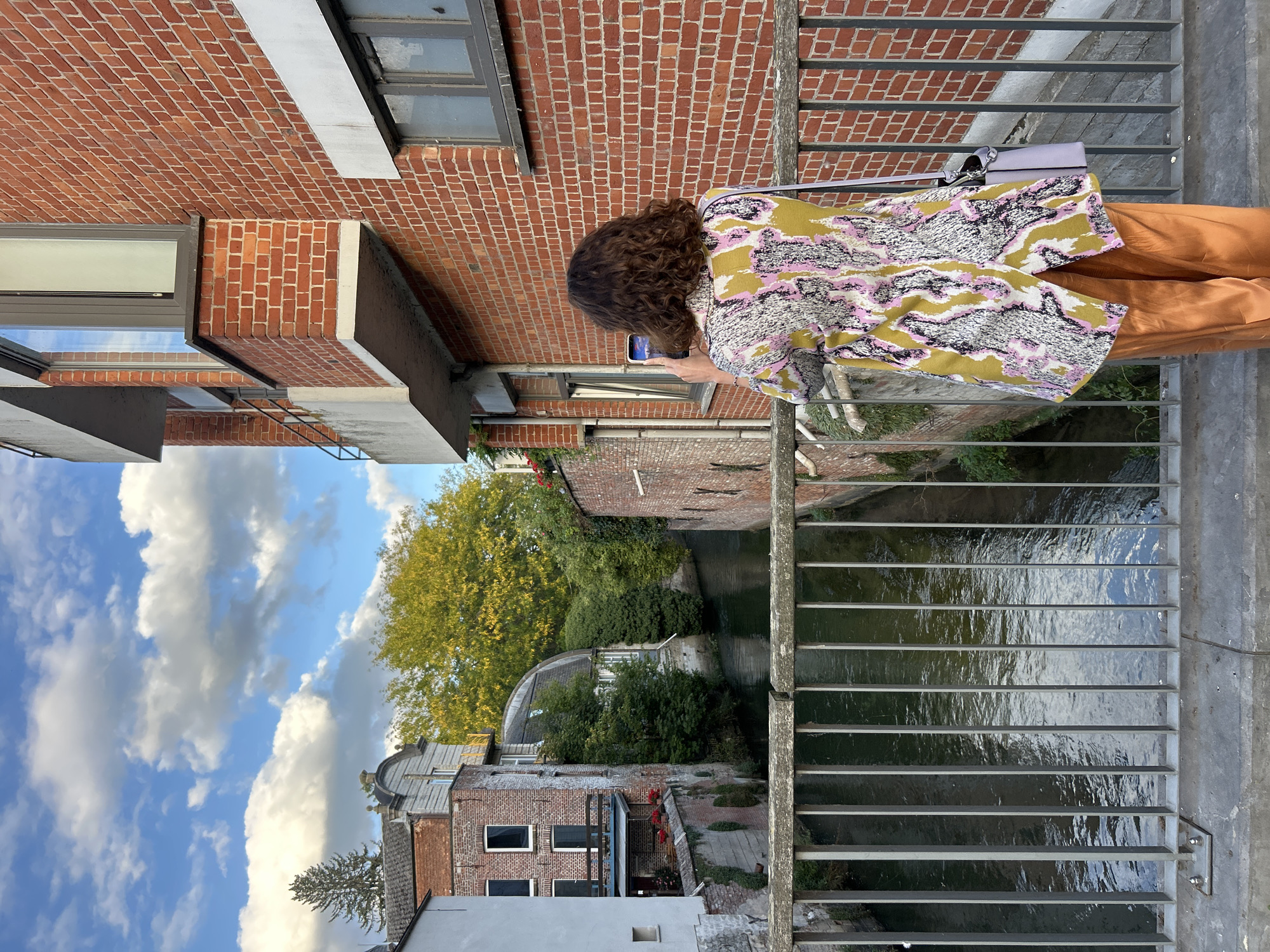 A woman with curly brown hair stands on a bridge in Leuven, Belgium, taking a photo with her phone. She is wearing a colorful, patterned jacket over orange satin pants. A light purple purse hangs from her shoulder. The bridge has a metal railing. To the left of the woman is a canal lined with old brick buildings and lush green trees. The sky above is blue with scattered white clouds. To the right of the woman is a modern brick building with a balcony and windows. The scene depicts a pleasant day in a picturesque part of the city.