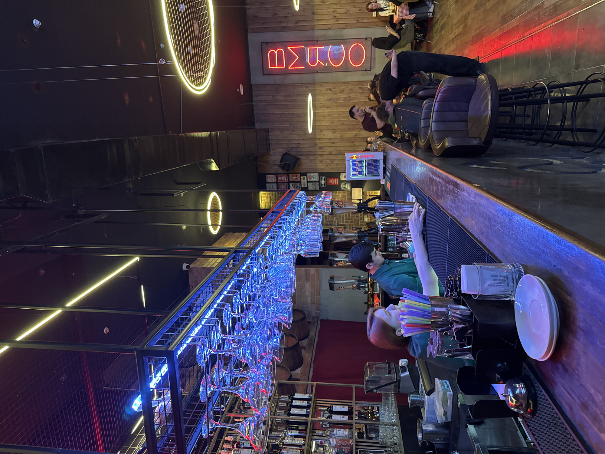 The image depicts the interior of a bar, likely in Tashkent, Uzbekistan, given the "Bmboo" neon sign (possibly indicating the bar's name). The scene is viewed from behind the bar, showcasing a bartender (likely female with reddish hair) and another person (likely male) working. Rows of glasses hang upside down from a blue-lit rack above the bar. Shelves behind the bar display various bottles of liquor. Customers are seated at the bar, some standing and conversing. The bar has a dark, modern aesthetic with neon lighting and wooden accents. The focal point is the bar counter with utensils, straws, and dishware.
