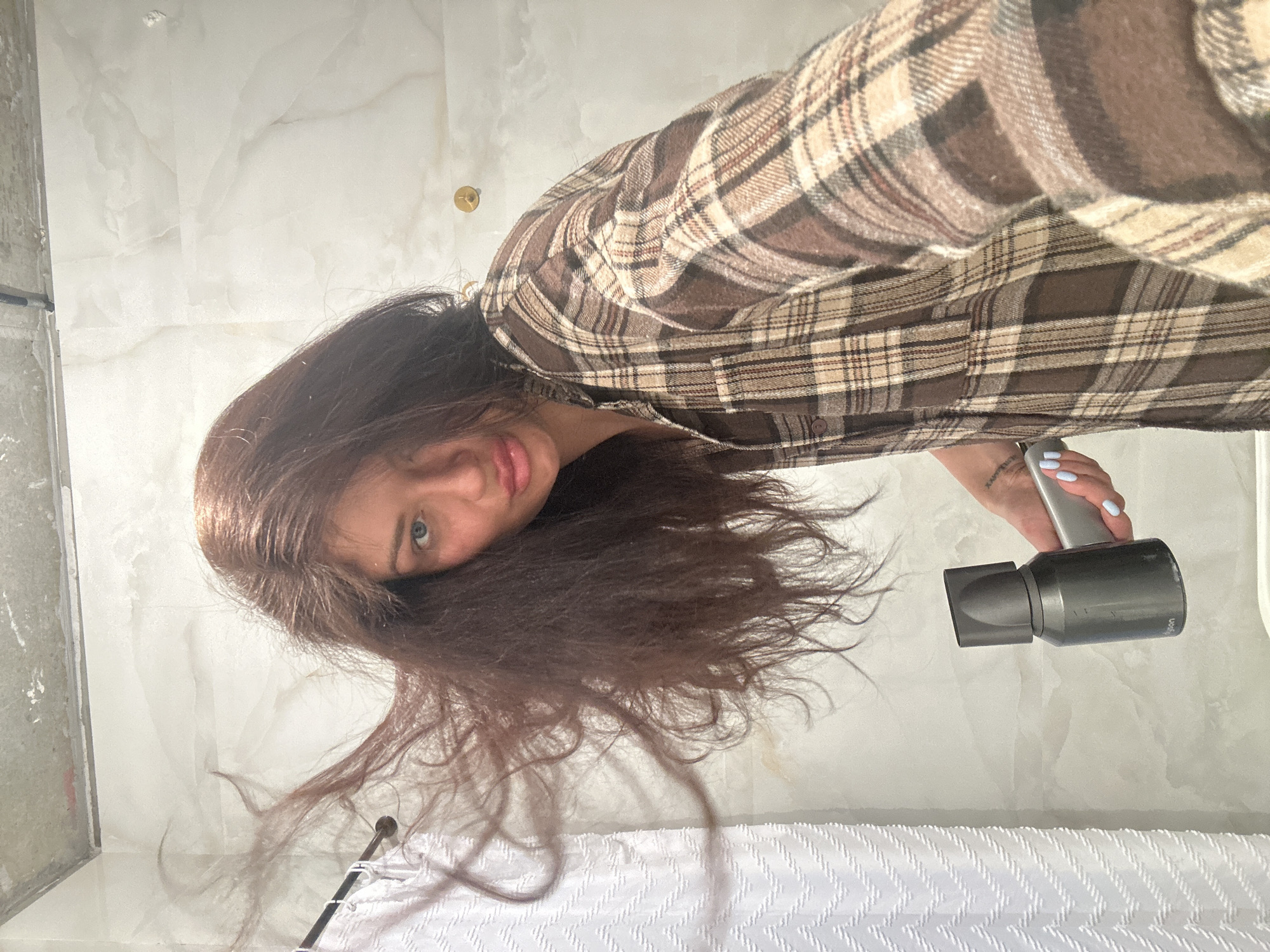 A woman is holding a Dyson Supersonic hair dryer in a bathroom. She is wearing a brown plaid shirt. The bathroom has white marble walls and a white floor. There is a small gold fixture on the wall. There is a white towel or blanket with a zig-zag pattern on the floor. The location is unknown and cannot be determined to be Moscow, Russia.