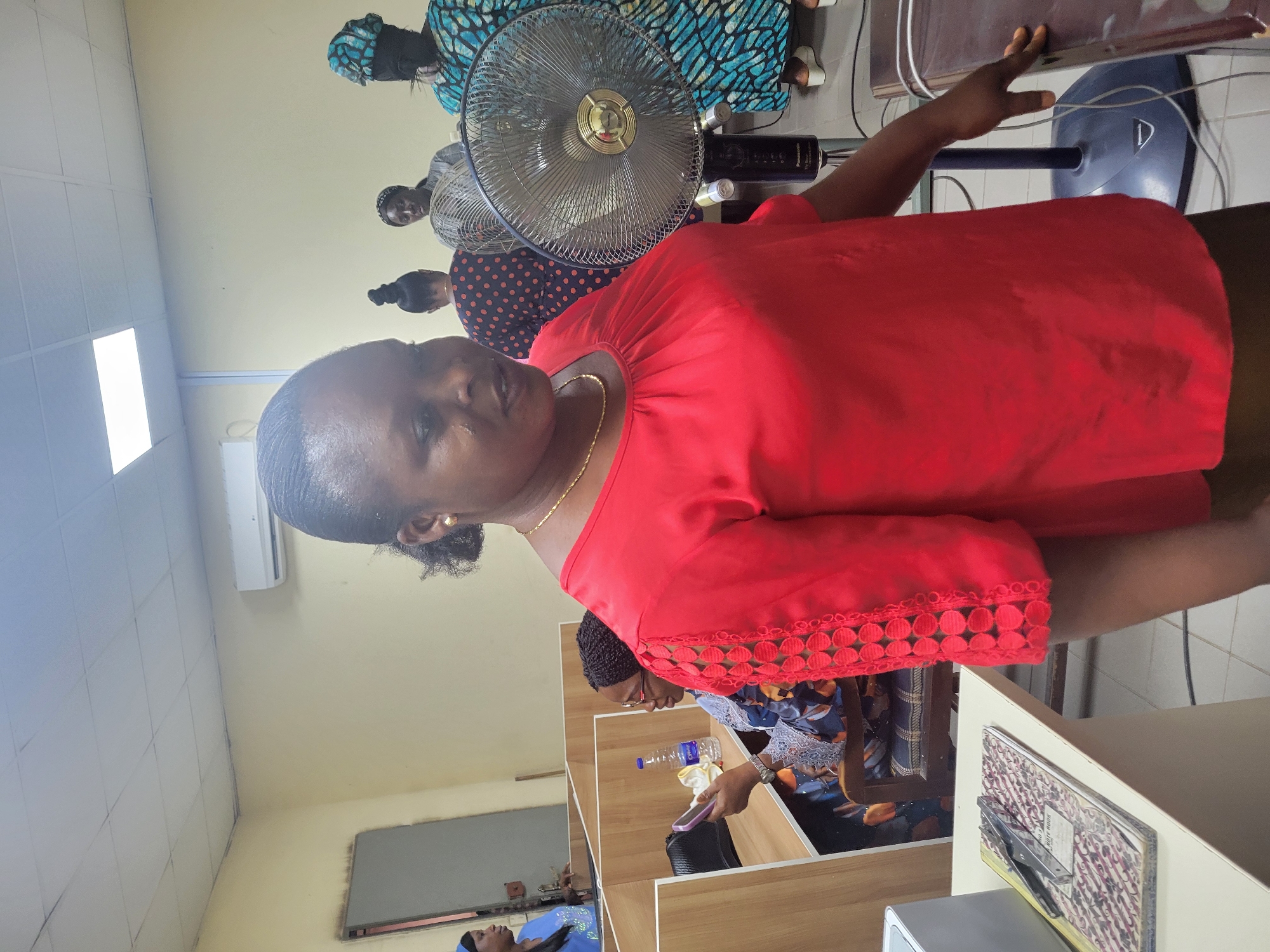 The image shows an indoor scene in what appears to be an office or meeting space in Abuja, Nigeria. A middle-aged woman with dark hair, wearing a red blouse with decorative trim on the sleeves and a gold necklace, is prominent in the foreground. Several other people, likely women, are in the background, some seated and wearing patterned clothing.
A metal standing fan is visible behind the woman in red. There are tables or desks with bottles of water and other personal belongings. The room has neutral-colored walls and a tiled ceiling with recessed lighting. A wall-mounted air conditioner is also visible.