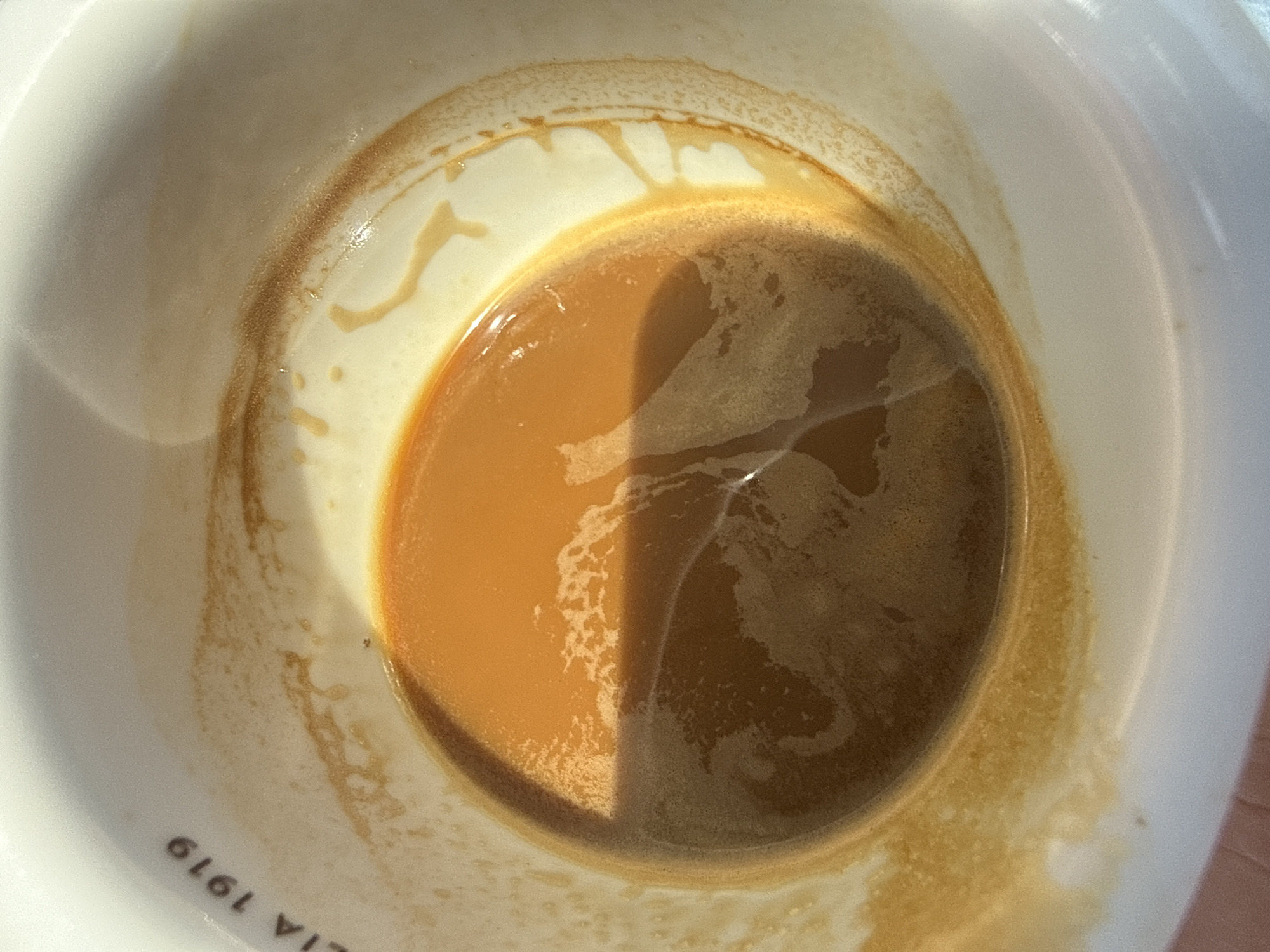 A close-up, overhead shot captures the inside of a white ceramic cup filled with amber-colored espresso. The espresso has a frothy layer on top, with some streaks of darker brown liquid mixed in. The cup's rim is visible, and it has faint brown stains from coffee residue. A partial shadow stretches across the surface of the espresso. The cup is branded with the text "ITALIA 1919" in dark, serif font on the upper left quadrant of the rim. The background is out of focus, but appears to be a bright, possibly outdoor setting. The scene is indoors, in the city of Unknown, Unknown.