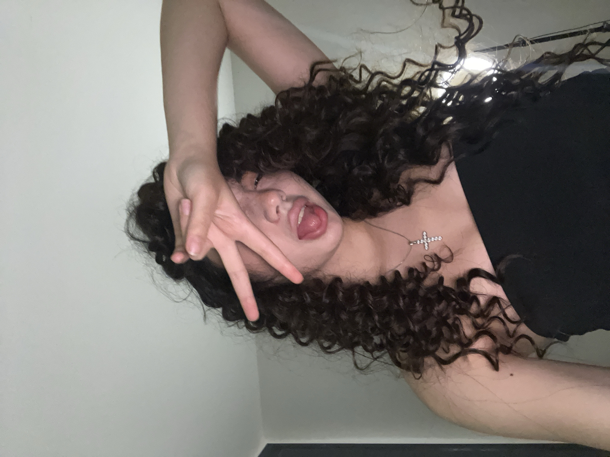 A young woman with long, dark, curly hair is shown from the chest up, indoors. She is wearing a black strapless top and a silver cross necklace. Her right hand is raised, obscuring part of her face, with her index and middle fingers extended in a peace sign. Her tongue is sticking out. The background appears to be a plain, light-colored wall, suggesting an interior setting. There is no information available about the location in Chotěšov, Czechia.