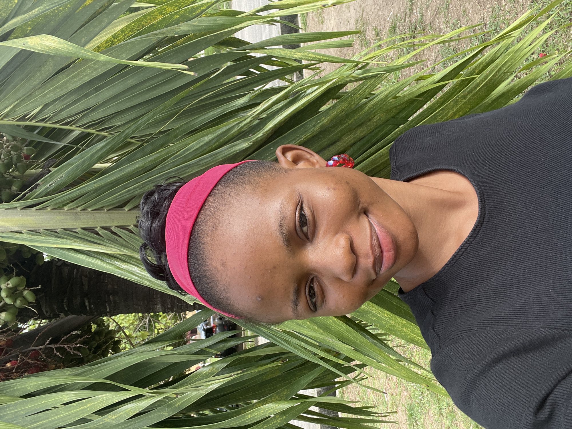 A woman with dark skin, wearing a black top and a pink headband, poses in front of a palm tree. She is smiling and has a red earring visible on her left ear. The background appears to be outdoors, possibly a park or garden in Ijanikin, Nigeria, given the presence of the palm tree. The immediate surroundings feature green foliage.