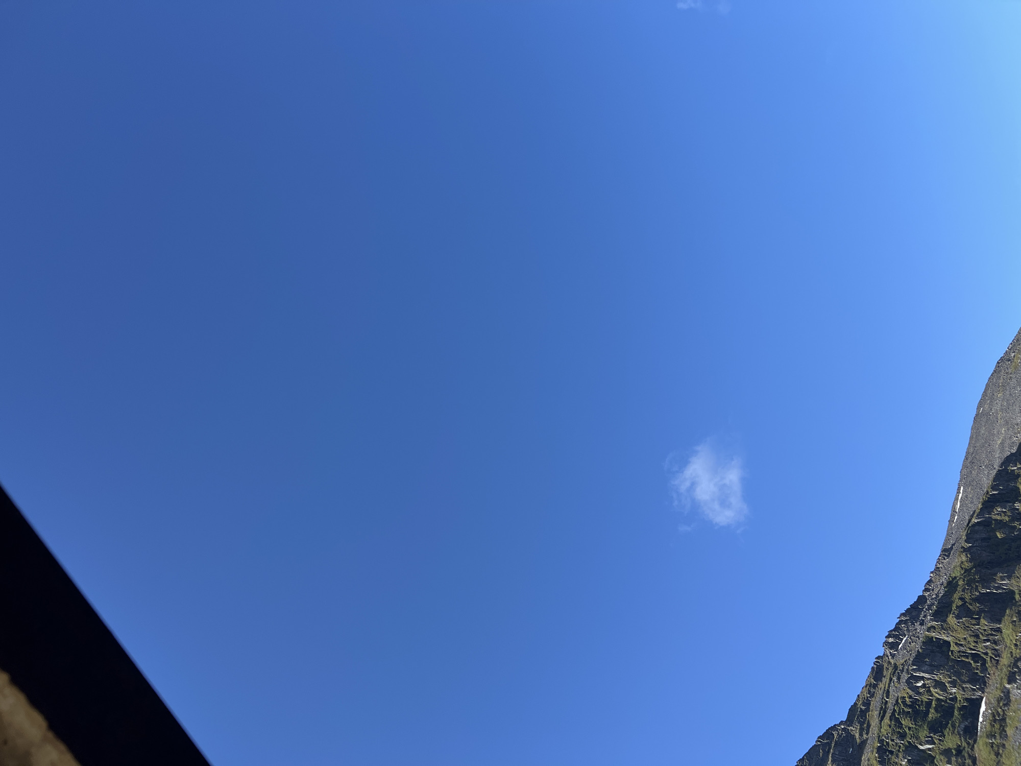 The image shows a clear blue sky with a single small cloud. In the lower left corner, a mountain range is visible. The mountains are steep and rocky, with patches of green vegetation and some snow. The top of the image is partially obscured by a dark, rough-textured object, possibly a building or overhang. The scene is likely outdoors in a mountainous region. Given the provided context of Kirovsk, Russia, this image may depict a view of the Khibiny Mountains.