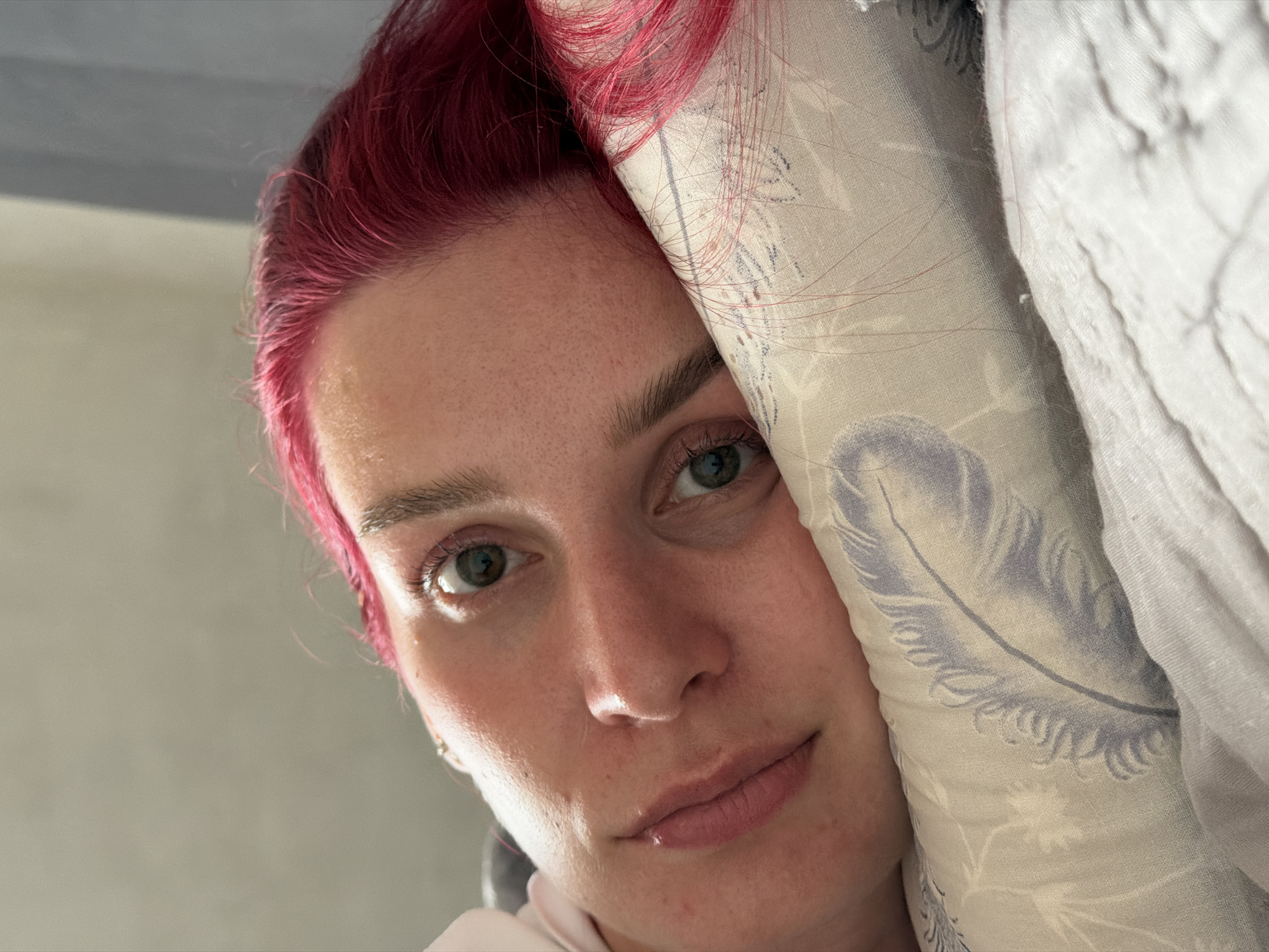A close-up portrait shows a woman with light skin, green eyes, and pink hair, partially obscured by a patterned pillow and folded white fabric. The scene is brightly lit. The location, Chaikovsky, Russia, is not discernible from the image.