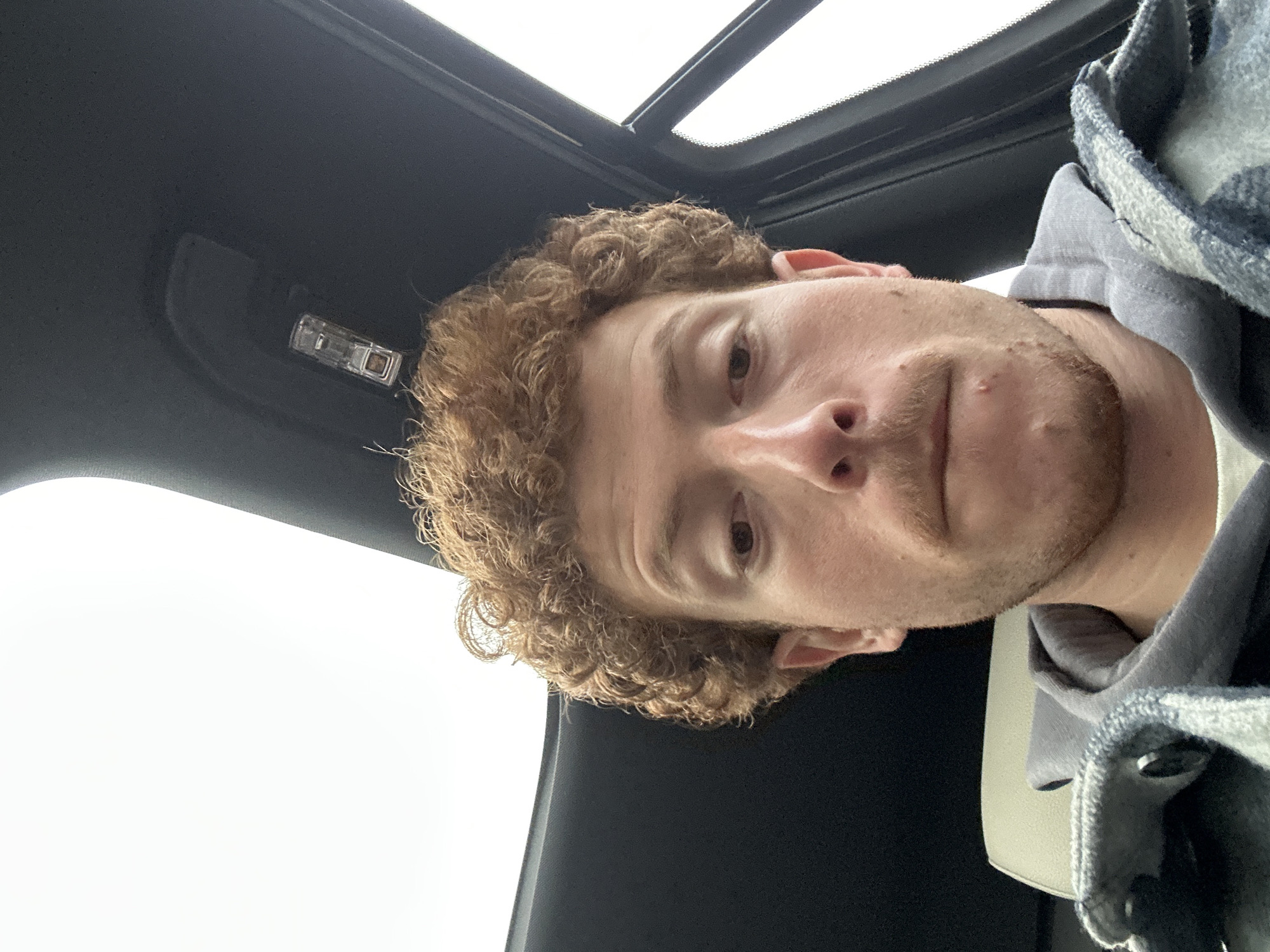 A close-up, upward-angled self-portrait shows a young man inside a vehicle. The man has fair skin, light brown curly hair, and light facial hair around his chin and upper lip. He is wearing a grey hooded sweatshirt underneath a blue, grey, and white plaid jacket. His expression is neutral as he looks directly at the camera.
The setting is the interior of a car, with a dark grey or black headliner visible around and above him. A bright, overcast sky is visible through a sunroof located in the upper right portion of the frame. An interior light fixture is present on the headliner towards the upper left. The diffused, bright light suggests it is daytime with cloudy or overcast weather. No specific details in the image indicate the precise location within Fort Lee, United States of America. There is no visible text in the media file.