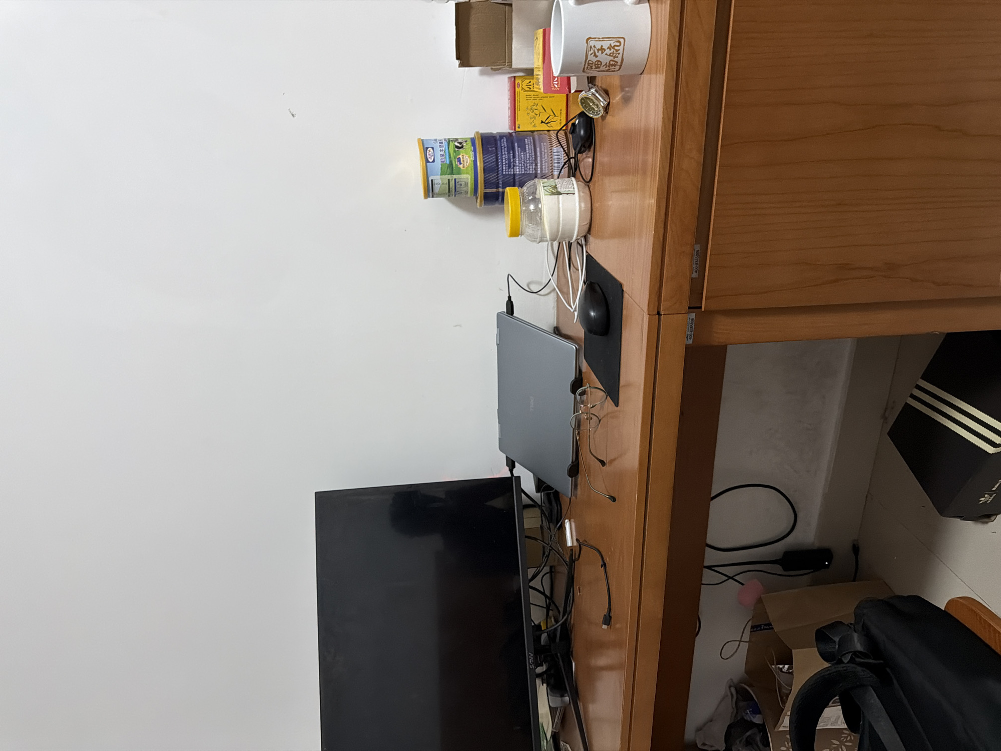 A cluttered desk stands against a white wall. On the desk are various objects, including a laptop, a computer mouse on a mousepad, a cup, cans, and boxes. Below the desk, there is a television and various bags. The specific location is Jiangning District, China.