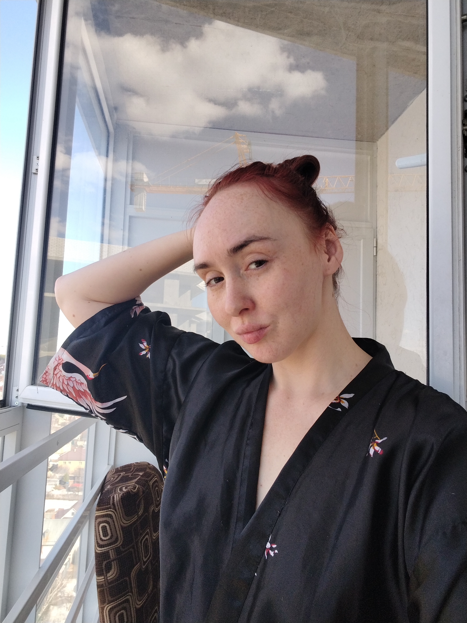 A woman with red hair pulled into a bun poses indoors by a window. She is wearing a black robe with a crane and floral pattern. The window offers a view of a city, featuring buildings and construction cranes. The image might be taken from a high-rise apartment in Izhevsk, Russia, given the urban landscape and cranes, reflecting potential construction activity in the city.