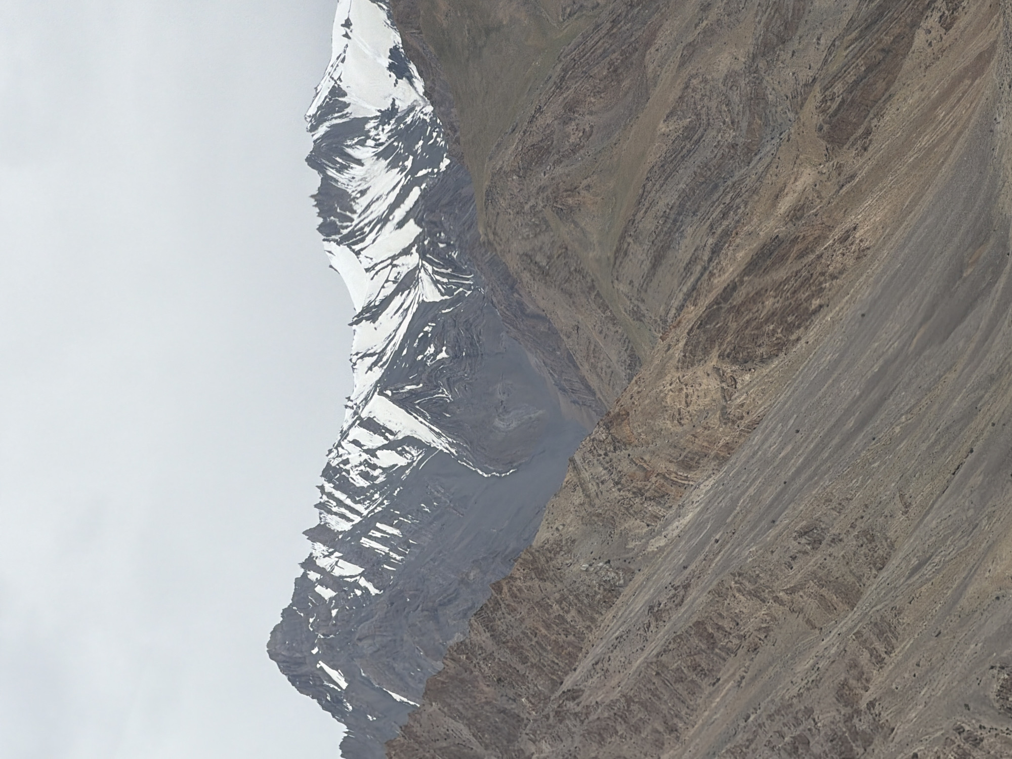 A mountainous landscape unfolds under a cloudy sky, with snow-capped peaks dominating the background. The foreground features rugged, layered slopes of rock and earth, displaying a range of earthy tones from brown to gray. The geology suggests a history of significant tectonic activity, evidenced by the folded and faulted strata. The scene is devoid of human presence or any man-made structures, indicating a remote and wild environment. The lighting is diffused due to the overcast conditions, casting a soft, even glow across the terrain and minimizing shadows. The scale of the mountains is immense, conveying a sense of grandeur and isolation. This is a natural scene, captured in the high-altitude region of Zangla, India, a place known for its stark, breathtaking mountain scenery.