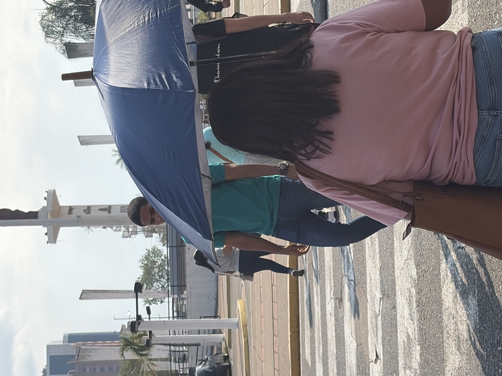 In Ciudad Guayana, Venezuela, the image captures a group of individuals crossing a street. A man wearing a turquoise shirt, jeans, and a dark-colored cap is under a blue umbrella. Nearby is a woman with dark hair, a pink shirt, blue jeans, and a brown purse. Other people are also walking on the crosswalk. In the background, buildings, trees, and street furniture, including lamp posts and decorative elements, are visible.