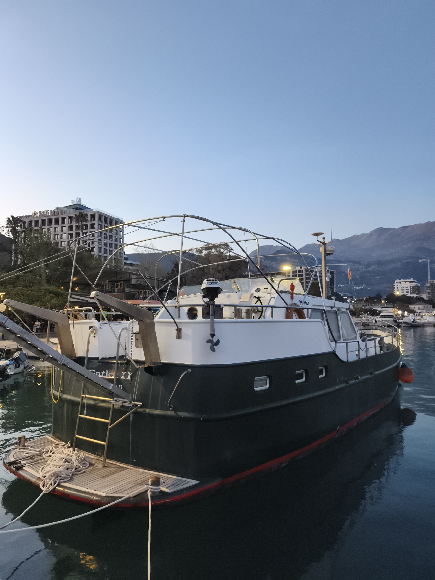 The image shows a boat docked at a marina in Budva, Montenegro. The boat, named "Catka II," is dark green and white with a red waterline. A metal frame surrounds the top. A wooden platform and ladder are on the stern. The sky is a clear gradient from light blue to pale orange, suggesting dusk or dawn. In the background, there is a partially constructed building, trees, and mountains. Other boats are visible in the harbor. The water is calm and reflects the boat and sky.