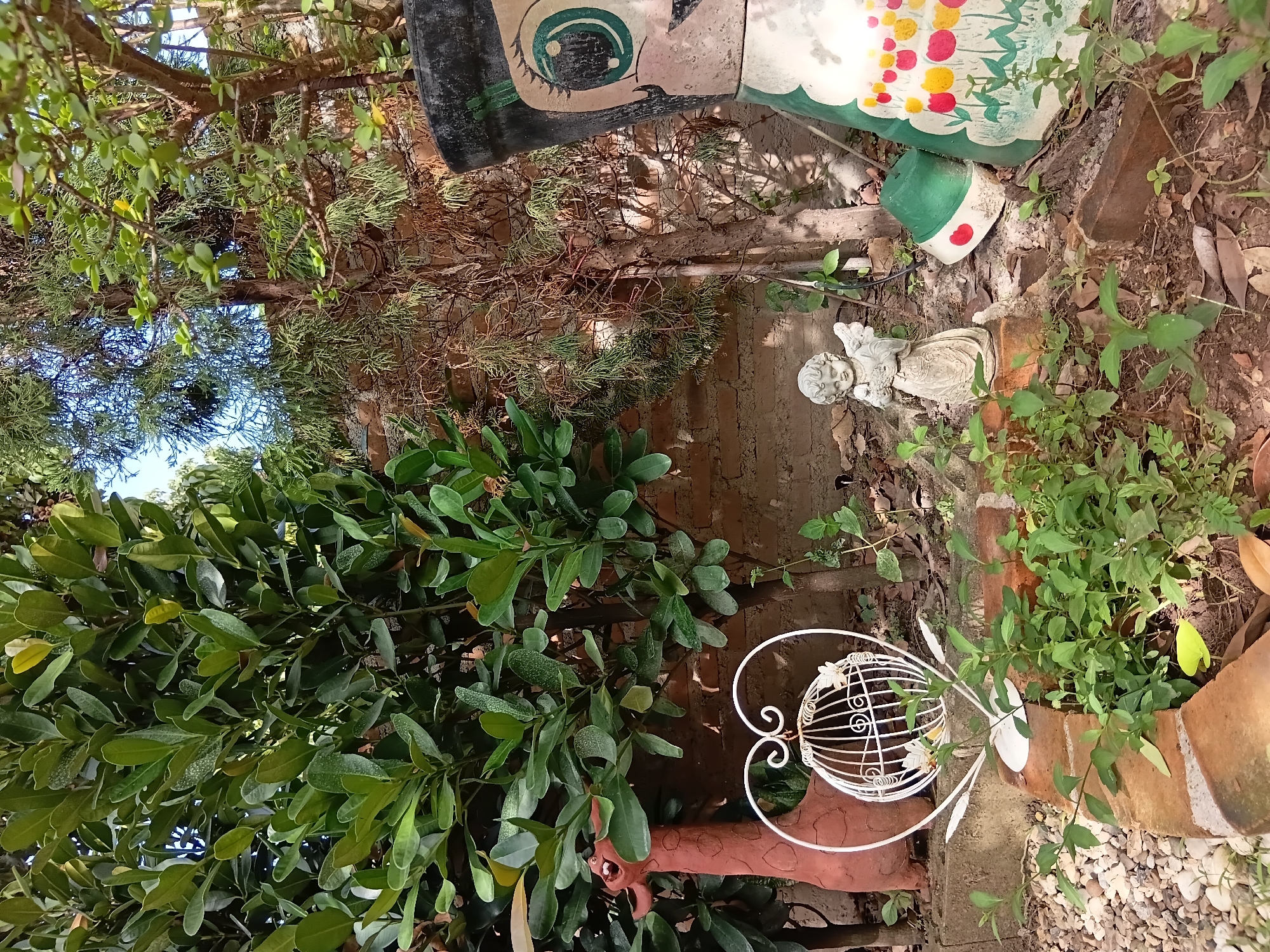 This image depicts an outdoor garden scene in Ban Tong, Thailand. The foreground features a small patch of green vegetation, with exposed brown soil and fallen leaves. Several bricks are arranged to form a border. To the left, a terracotta giraffe figurine stands next to a white heart-shaped decorative birdcage. In the background, a brick wall serves as a backdrop. A small, white angel statue is positioned in front of the wall, to the left of a green and white painted pot shaped like a figure's head, adorned with painted flowers. Lush green trees and foliage are visible throughout the scene, suggesting a well-established garden environment. The overall impression is one of a decorative, somewhat whimsical garden space.