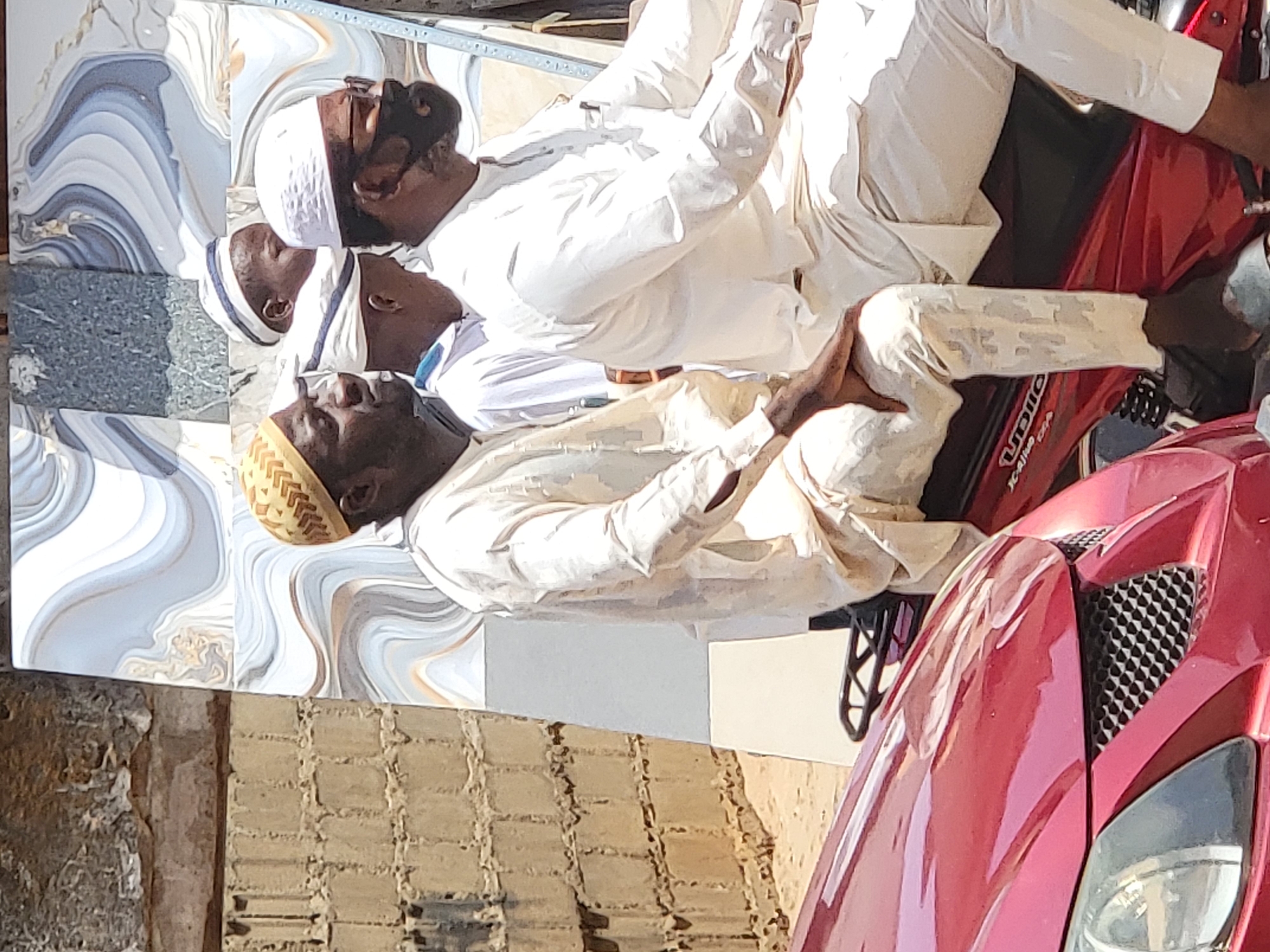 Three men, appearing to be of African descent, are captured in an outdoor setting in Maiburiji, Nigeria. They are all wearing traditional white or off-white garments, resembling thobes or kaftans, and head coverings, suggesting a Muslim cultural context.
One man, positioned on the far right, is sitting on a red motorcycle or scooter. He wears a white skullcap and a white garment, with a light brown belt visible. His right leg is bent, and his left leg is stretched forward.
In the center, another man is sitting or reclining, facing away from the camera, wearing a white striped cap and white attire. To his left, a third man is visible from the waist up, also wearing a white garment and a distinctive light brown, patterned kufi cap. He is looking upwards, away from the camera.
The background on the left features large, swirling patterned tiles in shades of blue, white, and brown, possibly decorative flooring or wall panels. Below these tiles, a rough, light-colored brick wall is visible, suggesting a building exterior or courtyard.
The scene is brightly lit by natural sunlight, indicating it is daytime. The men appear to be resting or engaged in casual conversation. On the side of the red motorcycle, faintly visible text includes the letters "UDIO" or "UBIDIO".