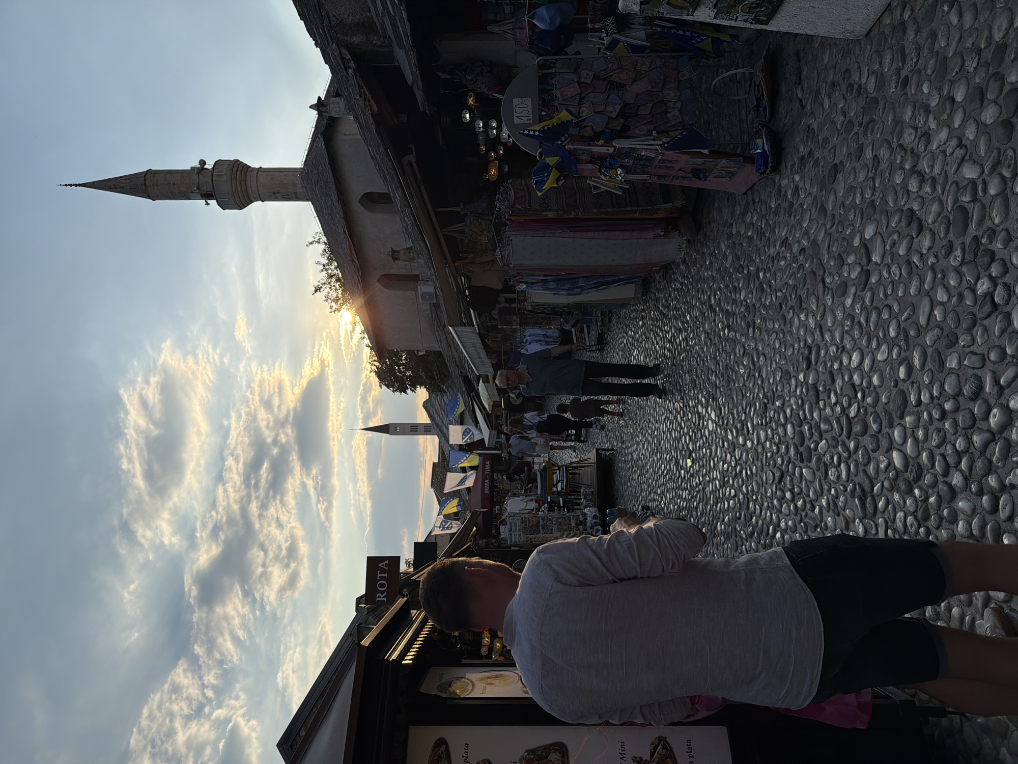 The image depicts a cobblestone street in the Old Town of Mostar, Bosnia and Herzegovina, during sunset. A man in a grey shirt and dark shorts stands in the foreground, facing away from the camera. The street is lined with shops selling various goods, including textiles, souvenirs, and lamps. The architecture features stone buildings with arched windows and tiled roofs. In the background, a minaret of a mosque pierces the sky, illuminated by the setting sun. Several people are visible walking along the street, and flags of Bosnia and Herzegovina are displayed on some stalls. The sky is partly cloudy, with the sun casting a warm glow over the scene.