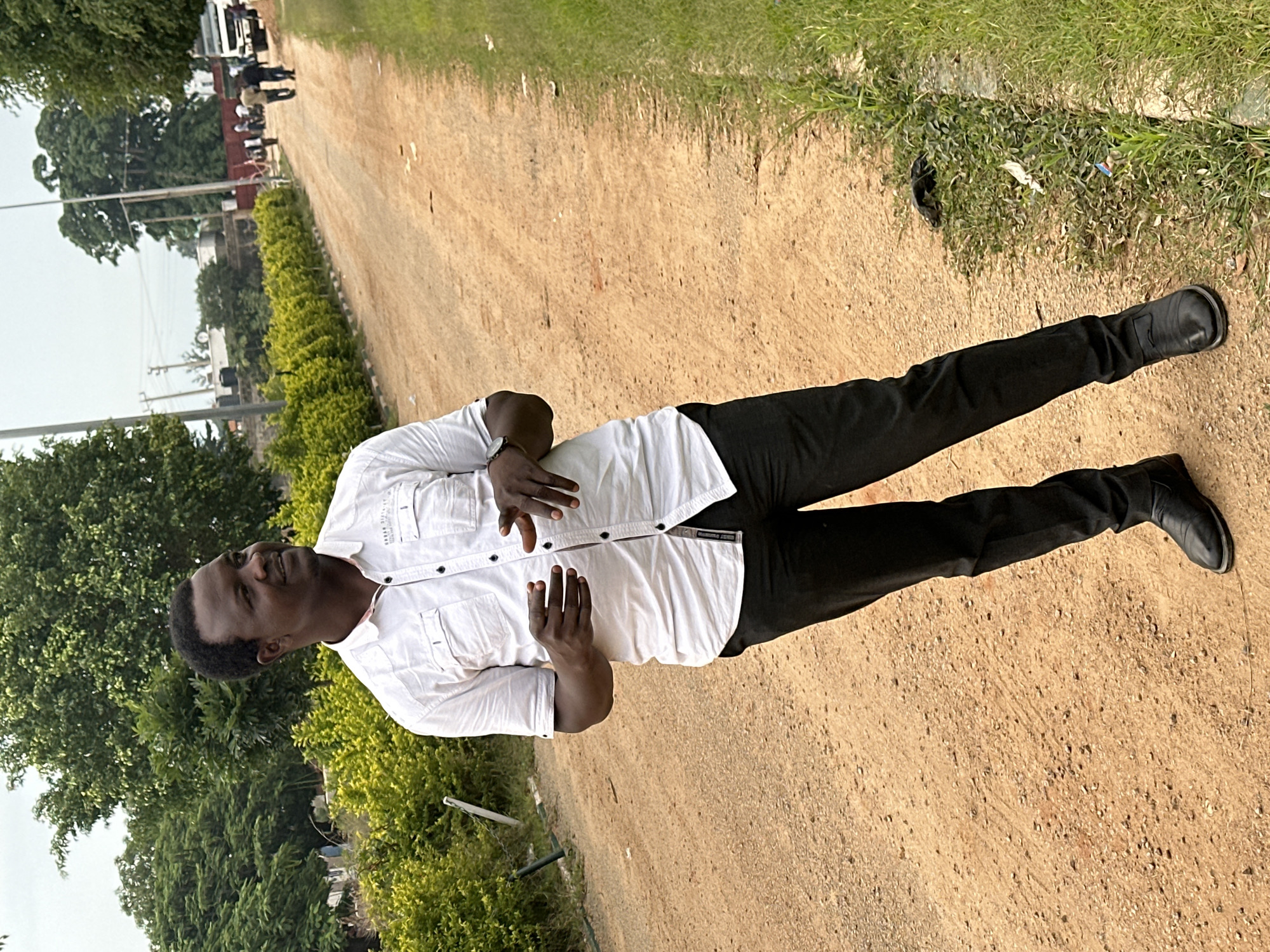 A medium shot captures a Black male standing on a dirt road in Dadi Riba, Nigeria. He is facing to his right, with his head turned towards the camera, offering a slight smile. He is wearing a short-sleeved white button-down shirt with two chest pockets and black pants. His right hand is slightly raised, with his fingers splayed, as if gesturing. He wears black dress shoes. The road is a light brown dirt path, flanked by green grass on the right and a line of manicured green bushes on the left. In the background, more trees and some buildings are visible. Several people are walking in the distance on the road, and a white truck is parked further down. The sky is overcast.