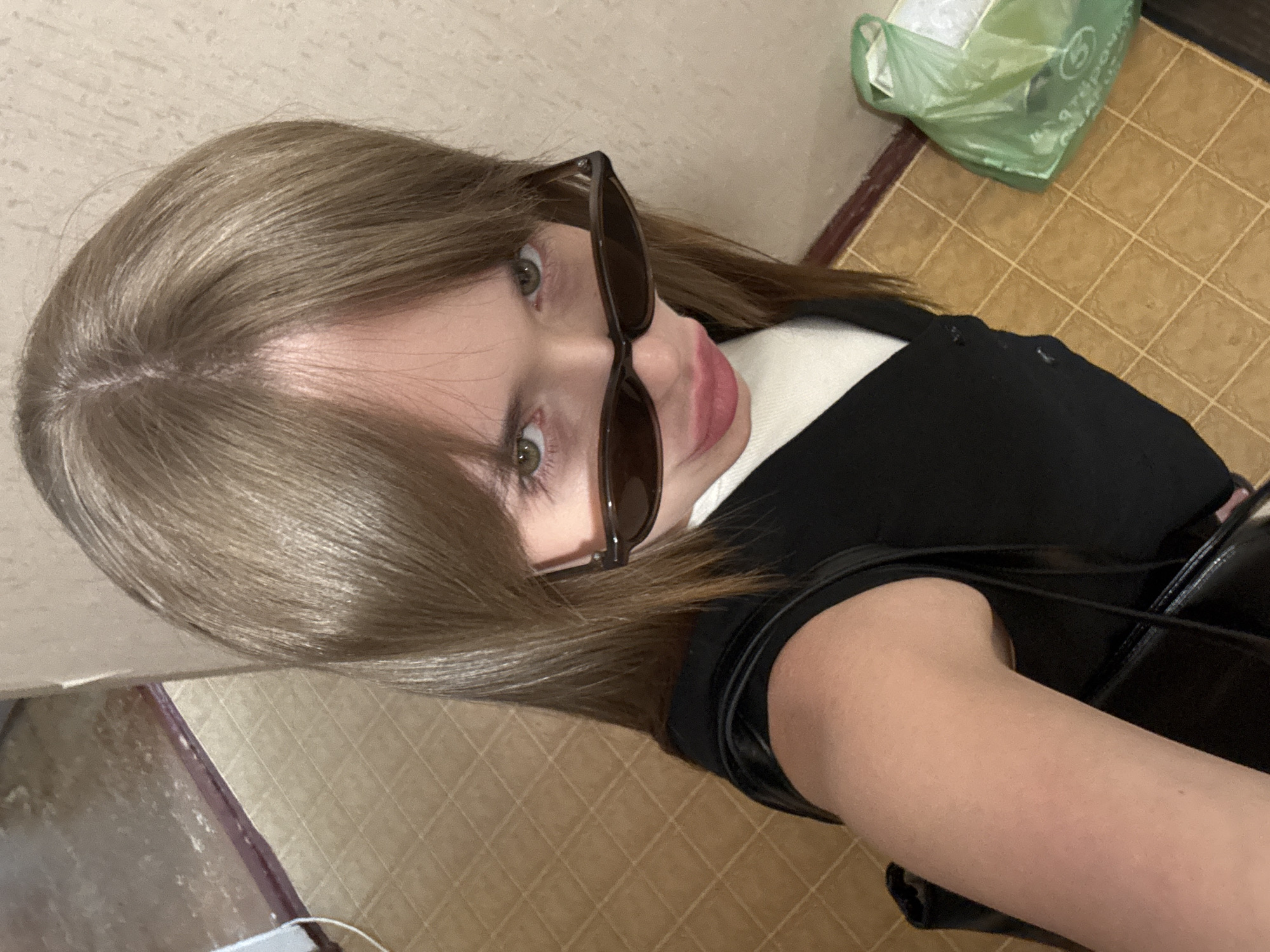 A person wearing sunglasses, a black top over a white collared shirt, and carrying a black purse is photographed from an upper angle. The individual has light brown hair. The location appears to be indoors, possibly a home, with tiled flooring and a green plastic bag visible in the background. The specific city of Chaikovsky, Russia, cannot be confirmed from the image alone.