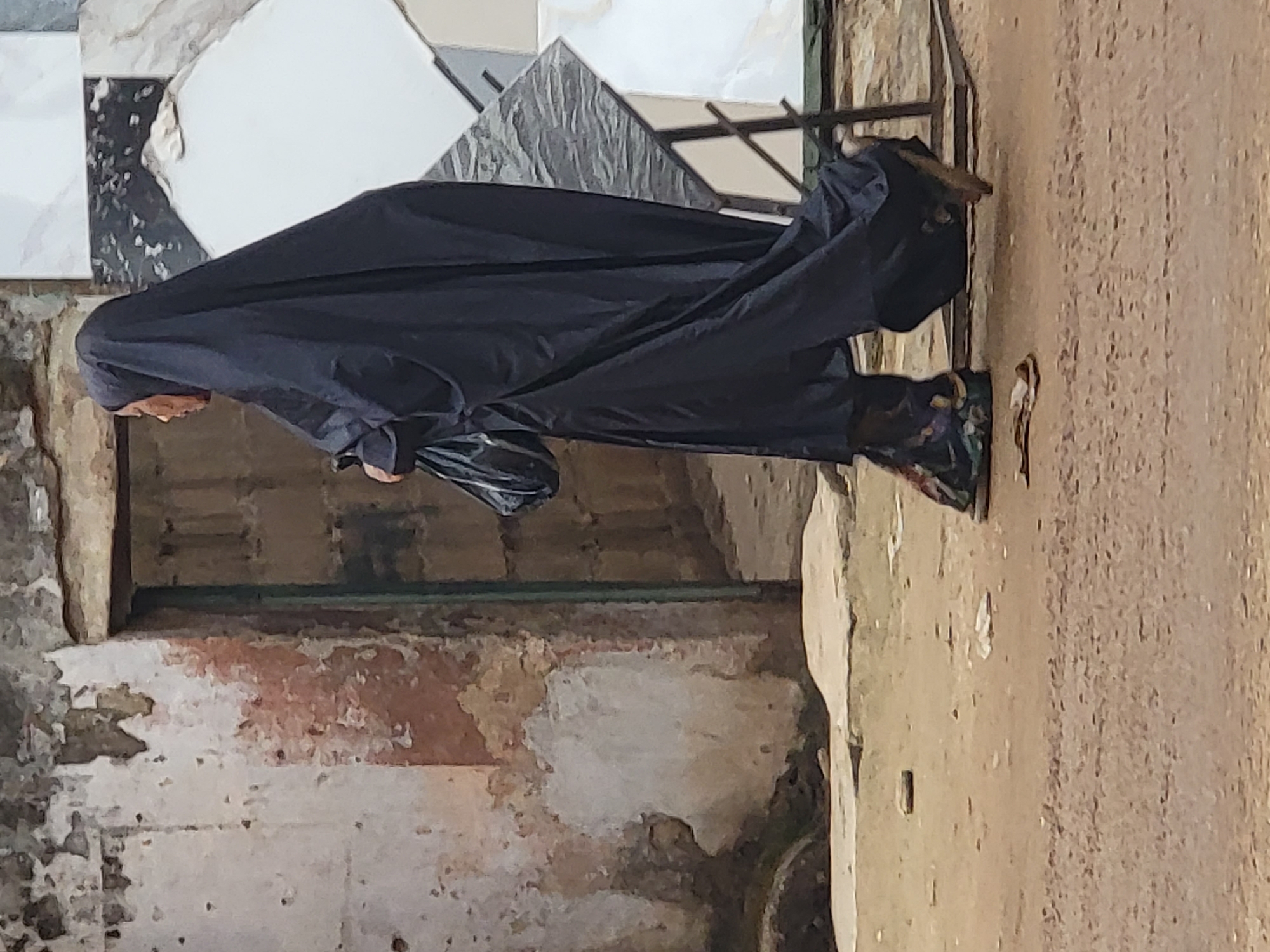 An outdoor, vertical full-shot captures a person standing or walking in what appears to be a street or commercial area in Maiburiji, Nigeria. The individual is dressed in a dark blue or black full-body garment, likely an abaya or similar attire, with a matching head covering that obscures most of the face, leaving only a glimpse of the chin and cheek visible. They are carrying a black bag, possibly plastic or fabric, in their left hand.
The background features large, square, light-colored tiles or slabs, possibly marble or ceramic, stacked against a wall or displayed for sale. Some dark metal shelving or a framework is visible behind the person's right side. The ground beneath the person is rough and uneven, suggesting an unpaved or poorly maintained surface. To the person's right, a textured, light-brown wall shows signs of wear, with patches of flaking plaster or paint, and rough material underneath. The overall lighting indicates it is daytime. The person's brightly patterned shoes offer a contrast to their monochrome attire. No specific weather conditions are discernible, and no text is visible in the image. The setting suggests an everyday scene in a bustling, possibly commercial, outdoor environment.