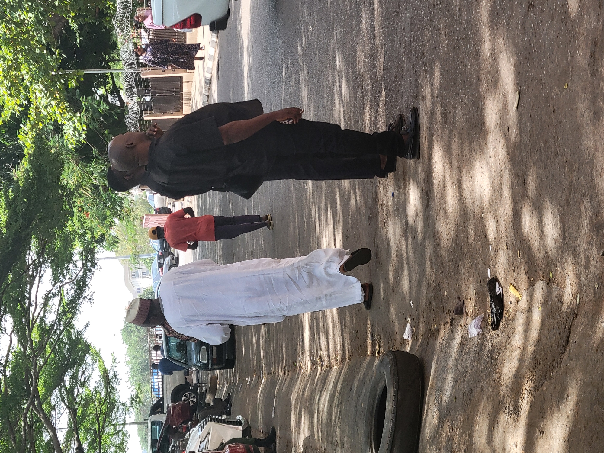 This sunlit outdoor photograph captures a street scene, characteristic of an urban or semi-urban setting, consistent with Abuja, Nigeria. The ground, a mix of dirt and possibly degraded pavement, is dappled with strong shadows cast by overhead trees, indicating a sunny day, likely midday or early afternoon.
In the foreground, a prominent figure, a man dressed in a black long-sleeved shirt, black trousers, and dark shoes, stands facing slightly left with his head tilted upwards. He is bald and appears to be holding a small object, possibly a phone or cigarette, in his right hand. Partially obscured behind him is another individual in dark attire. To his right, a man in traditional white clothing, including a flowing white robe and a patterned cap, stands with his back mostly to the camera, wearing sandals. Further in the background, a person in a reddish-orange top and dark bottoms is also visible, along with other distant figures.
The left side of the frame is lined with vibrant green trees, providing ample shade. In the mid-ground, behind the man in black, a fence topped with razor wire is visible. Several vehicles, including cars and possibly a truck, are present in the background, some parked and others partially obscured. The ground exhibits some litter, including plastic bags and other debris, while a large, worn-out tire rests in the lower right corner. The scene depicts a casual environment with people standing around, suggesting everyday activity rather than a specific event. No discernible text is visible within the image.