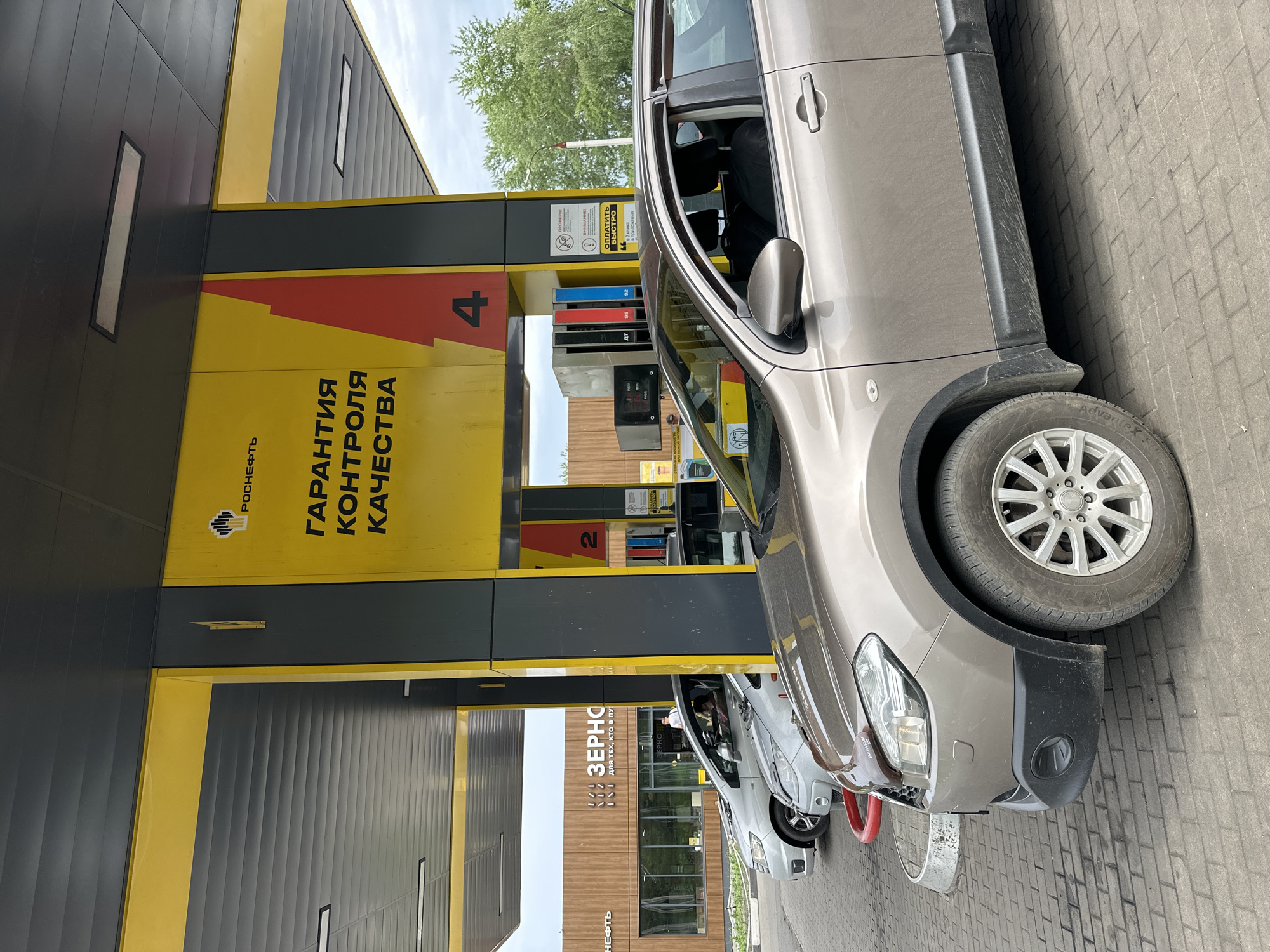 The image shows a gas station with cars lined up to refuel. A prominent yellow and black sign displays the "Rosneft" logo and text in Russian, including "Гарантия контроля качества" (Quality control guarantee) and the number "4". Another sign on the gas pump has the number "2". In the background, a building with "Зерно" (Grain) written on it is visible. A few people are present, including drivers in their cars and a person standing near the building. The scene appears to be a typical gas station environment, with cars, pumps, and associated signage. The location is identified as Slyudyanka, Russia.