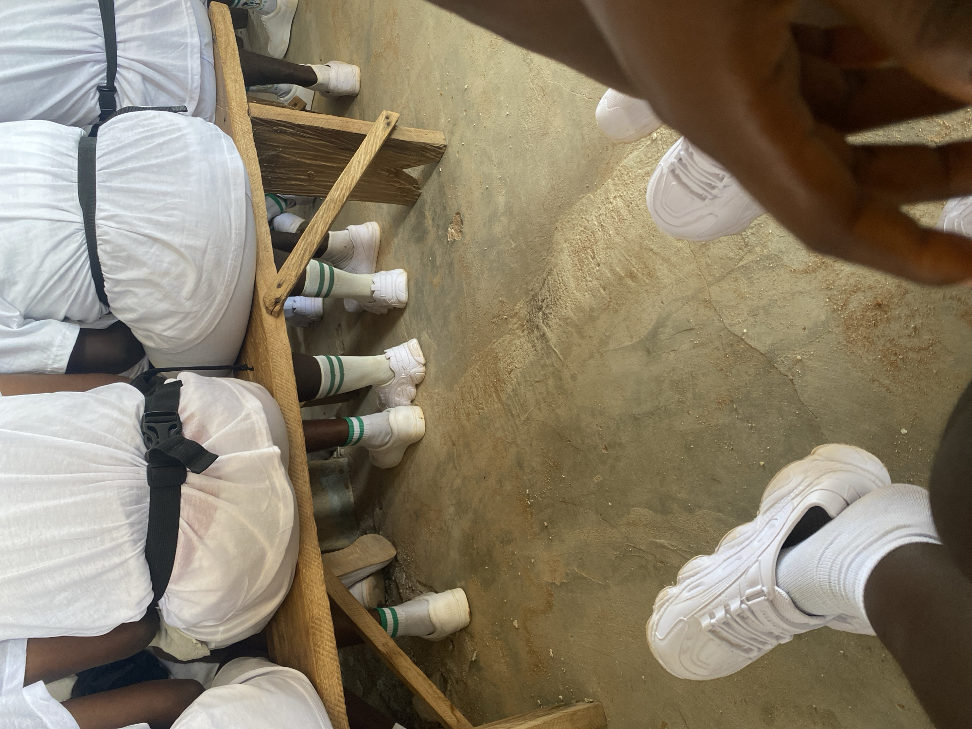 The image shows a group of people, likely children or young adults, sitting on a wooden bench outdoors, with a concrete floor. They are all wearing white uniforms, including white t-shirts, white shorts, white socks with green stripes, and white sneakers. Some individuals have black belts cinched around their waists over their white uniforms. The bench appears to be made of rough-hewn wood and is positioned on a dusty, uneven concrete surface. The background is out of focus but suggests an outdoor setting with natural light. The scene appears to be a gathering or waiting area, possibly at a school or community event. The people are not interacting with each other in the frame, and their attention seems to be directed forward or downward. The overall impression is one of order and uniformity due to their matching attire. The specific location in Jimeta, Nigeria, cannot be determined from the image alone, but the context of the uniforms and the setting might suggest a school or organized group activity common in such regions.