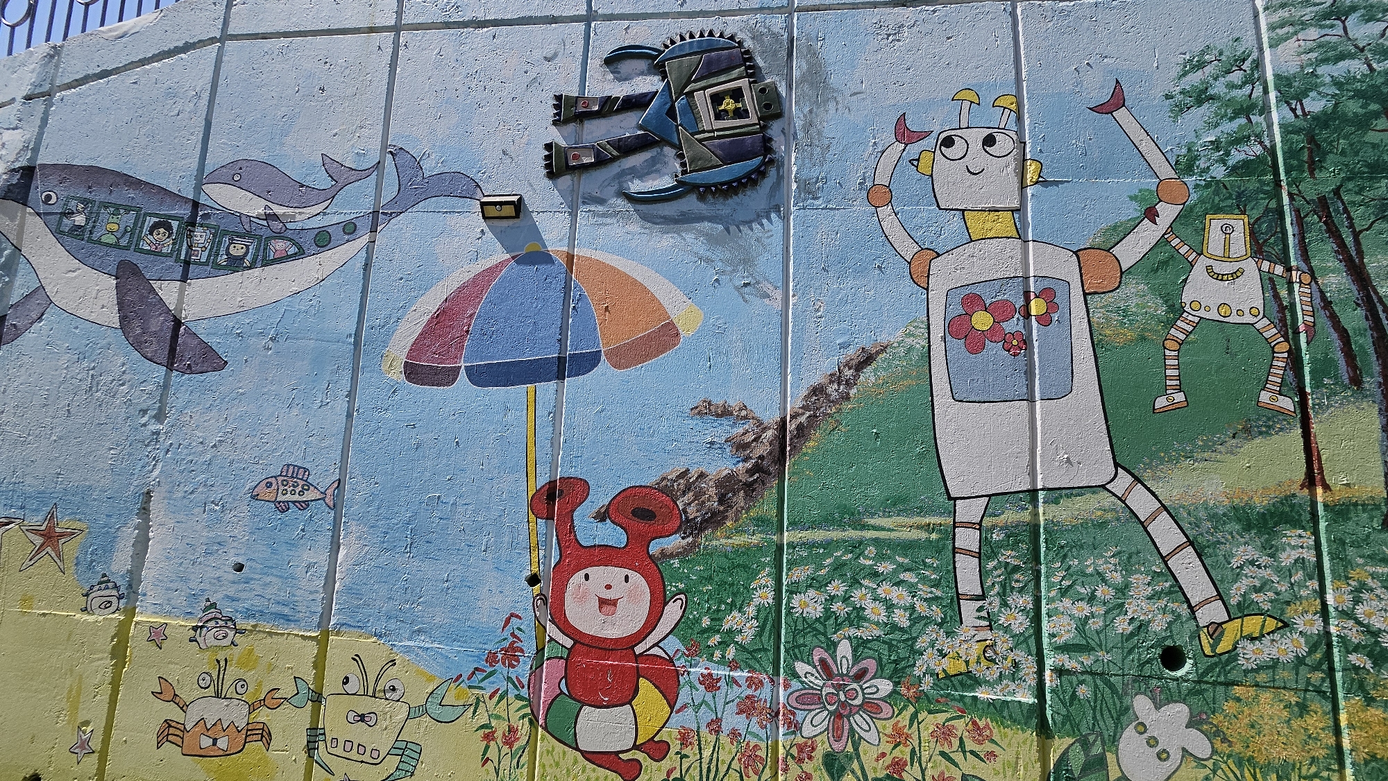 A mural painted on a segmented wall in Ulsan, South Korea, depicting an underwater scene with a whale containing figures, a fish, and crabs at the bottom; a red bug-like character under an umbrella, a robot standing among flowers, and another robot in the background among trees, with a geometric robotic figure attached above. The painting shows signs of wear and weathering.
