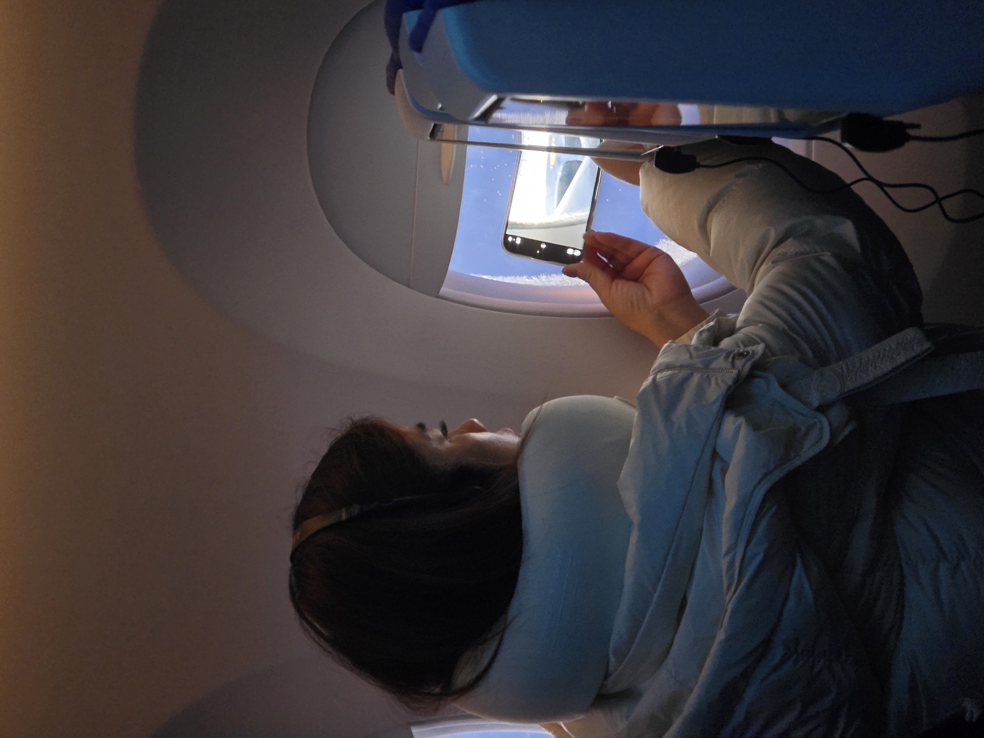 The image shows a person inside an airplane cabin, viewed from a slightly upward perspective. The subject, likely female, with dark hair, is seen in profile, looking towards a brightly lit oval airplane window. She is dressed in a light-colored top or jacket, with a light-colored blanket or cushion draped around her.
Her main activity is holding a black smartphone up to the airplane window, seemingly capturing a photograph or video of the outside view. The phone's screen clearly reflects and displays the scene it is capturing: a bright blue sky with white elements, which appear to be clouds or a part of the aircraft's wing or engine. The person's right hand is visible holding the phone.
The setting is distinctly an airplane, characterized by the unique window shape and cabin structure. Above the window, a blue and white panel or overhead compartment is visible. Black cables are seen hanging in the upper right quadrant of the image. The ambient light inside the cabin appears softer and dimmer than the bright daylight visible through the window. There is no discernible text visible on the smartphone screen apart from standard camera interface icons. The scene suggests a daytime flight where the passenger is documenting the aerial view.