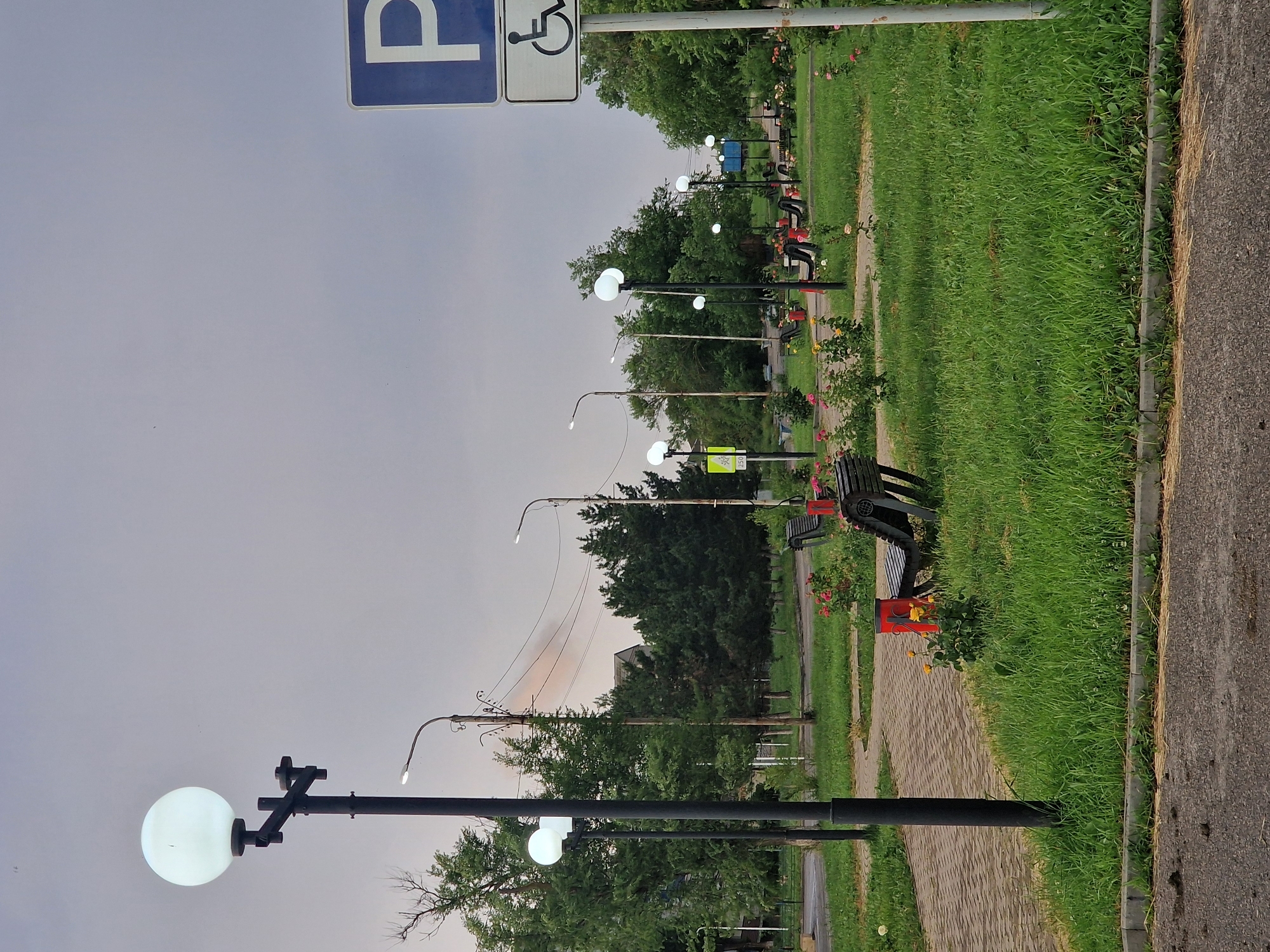 The image depicts a public park area in the city of Kursavka, Russia, under an overcast sky. A paved walkway, made of interlocking bricks, winds through a well-maintained grassy area. Several lampposts with spherical, illuminated globes line the walkway, providing light. Benches are strategically placed along the path, some with visible occupants (not clearly discernible as people). In the background, a dense line of trees forms a natural boundary, with glimpses of buildings beyond. A parking sign with a blue "P" and a wheelchair accessibility symbol is prominently displayed on the right side of the frame, indicating a designated parking area. Several rose bushes with pink and yellow blooms are scattered throughout the park, adding a touch of color to the scene. The overall impression is one of a tranquil, urban green space.