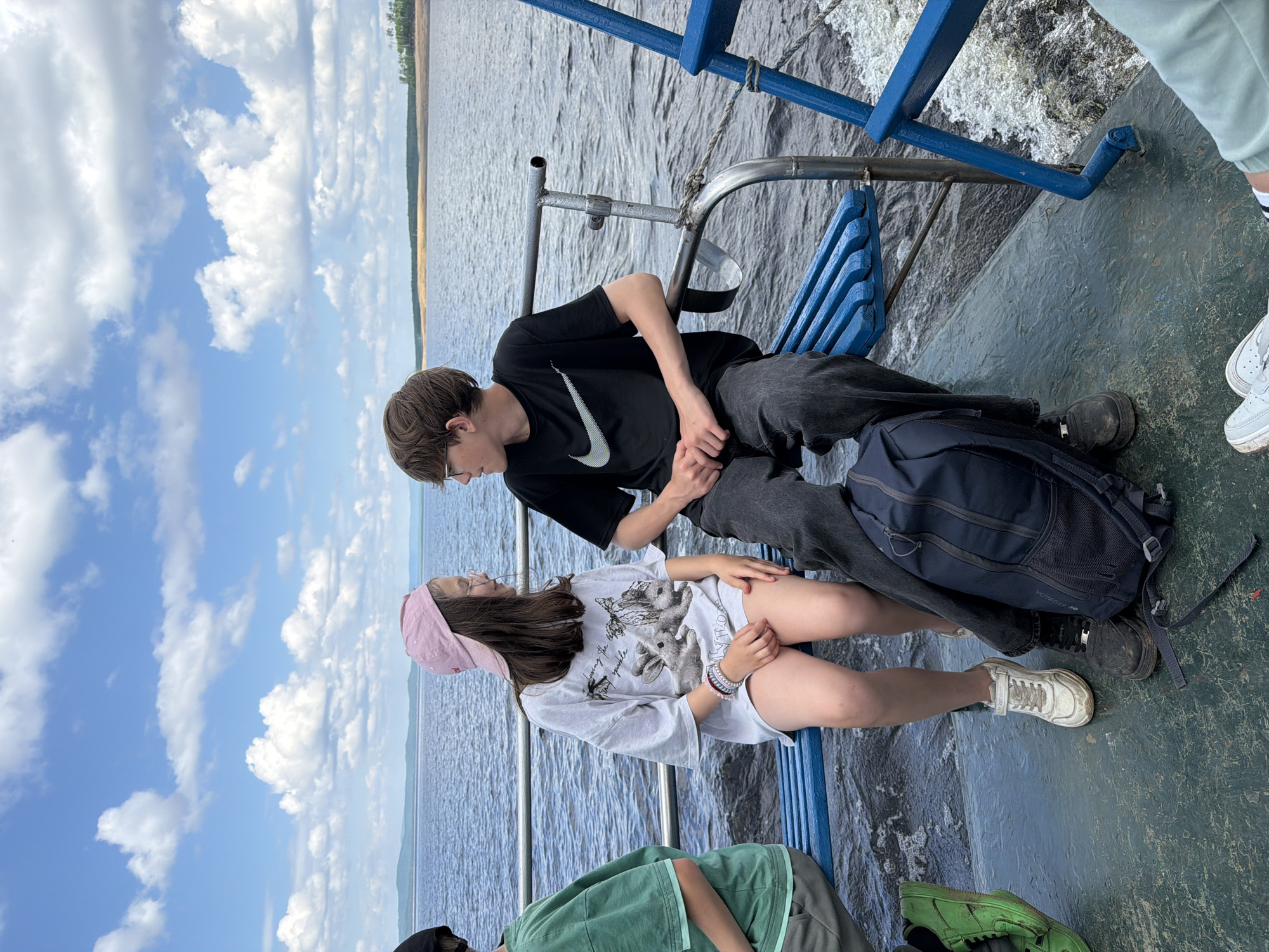 Two children, a girl and a boy, are seated on a blue bench on a boat, looking at each other. The girl, wearing a pink baseball cap and a white t-shirt with a graphic print, has bracelets on her wrist. The boy, in a black t-shirt with a Nike logo, is wearing dark pants. A navy blue backpack is placed on the deck between them. The boat is on a body of water under a sky filled with white, puffy clouds. The background shows a distant shoreline with trees and hills. The deck of the boat is green and appears worn. To the right, a blue railing is visible, with white frothy water from the boat's movement.