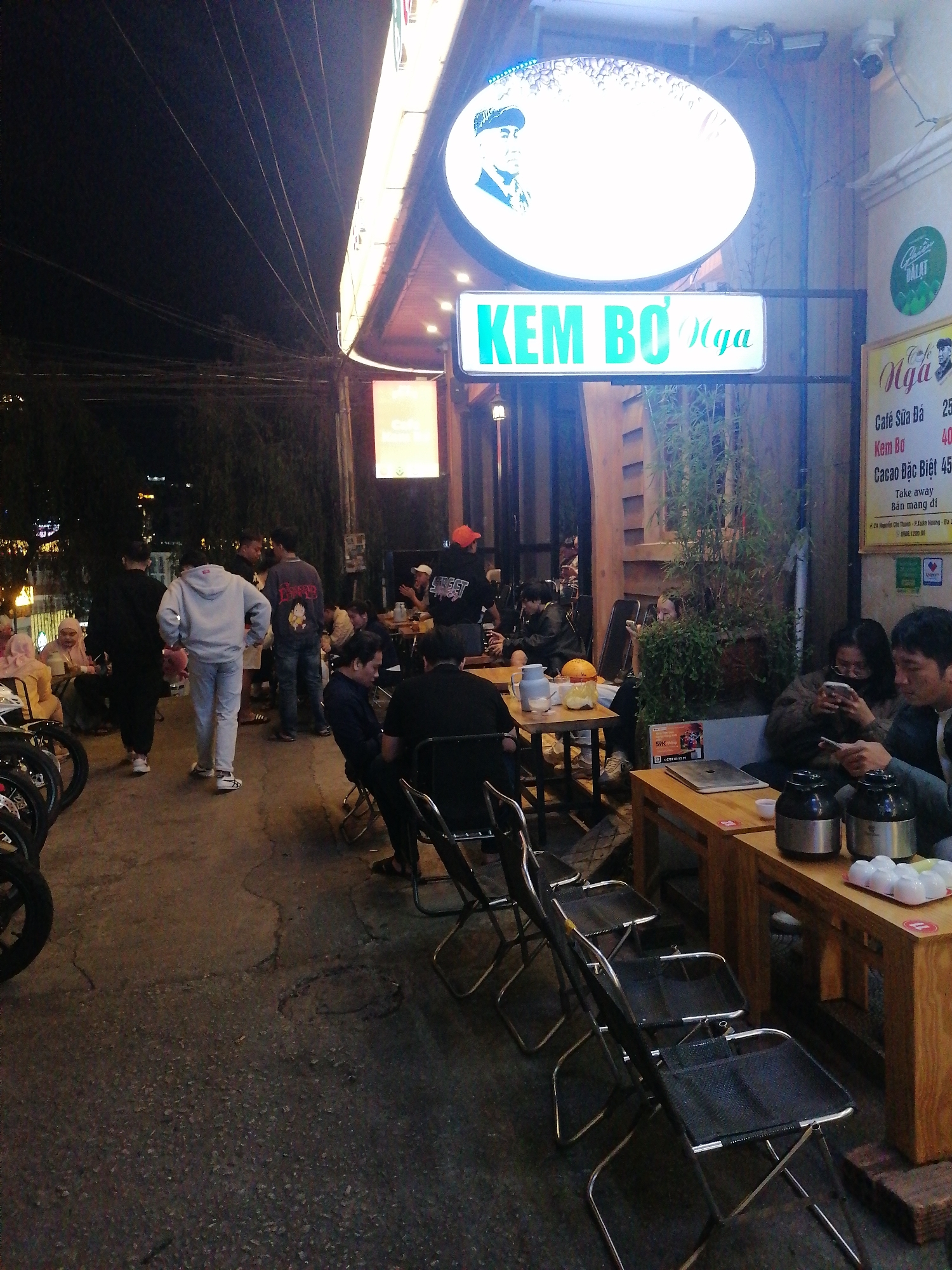 The image shows the outdoor area of "Kem Bo Nga," a dessert and coffee shop in Da Lat, Vietnam, at night. Several people are seated at wooden tables, some looking at their phones, others engaged in conversation. There are metal folding chairs placed around the tables. In the background, other people are walking by. Motorcycles are parked on the left side of the image. The shop's signage, illuminated, displays "KEM BO Nga" and a circular sign with a portrait. A menu board shows prices for Cafe Sua Da (25), Kem Bo (40), and Cacao Dac Biet (45). Steam kettles and small white cups are on one of the tables. The street is concrete and slightly uneven. The environment appears to be a popular, bustling area in the city.