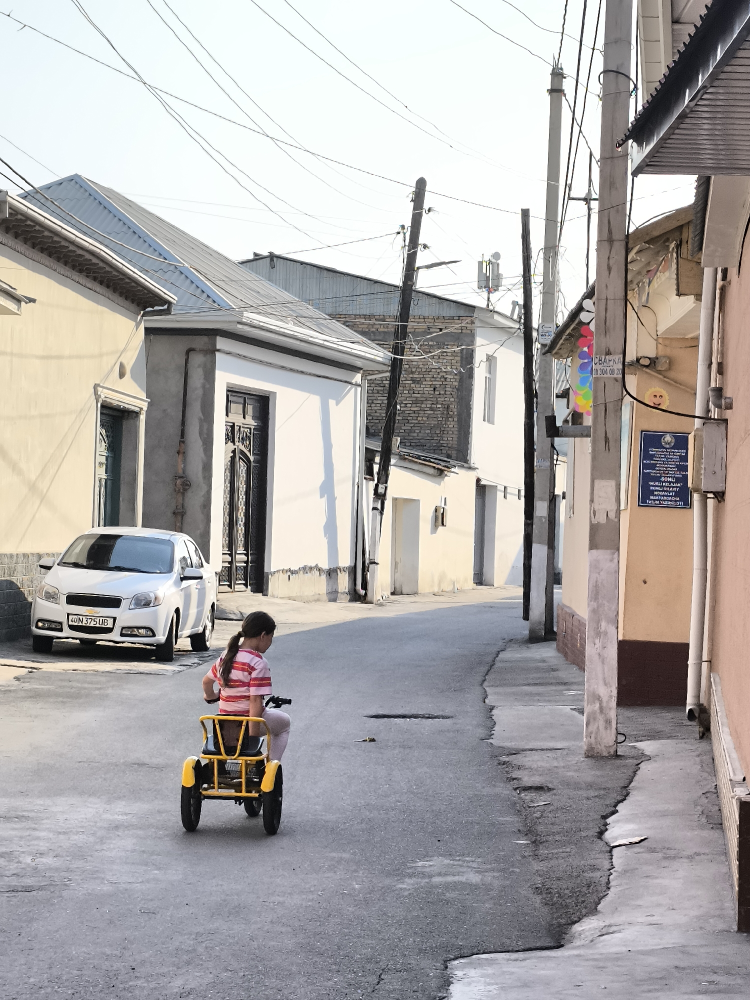 In the city of Kokand, Uzbekistan, a young girl with dark hair pulled back in a ponytail rides a yellow pedal-powered vehicle on a narrow, paved street. She is wearing a pink and white striped shirt and light-colored pants. The street is lined with buildings, some with white facades and others in shades of beige and peach. A white sedan is parked on the left side of the street, and various utility poles and overhead wires crisscross the sky. A sign with writing in Cyrillic script is mounted on a pole on the right side of the street. The lighting suggests it is daytime, possibly late afternoon, with shadows cast by the buildings.